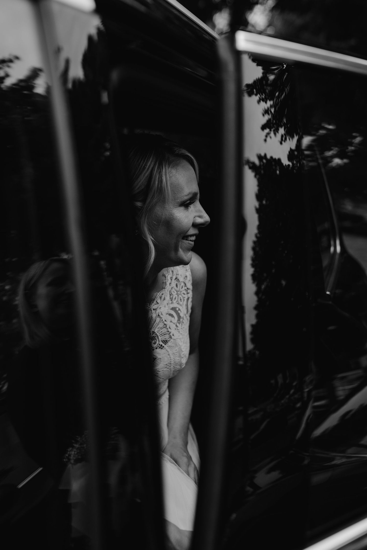A black and white portrait of someone in a white lace dress smiling through foliage and shadows.