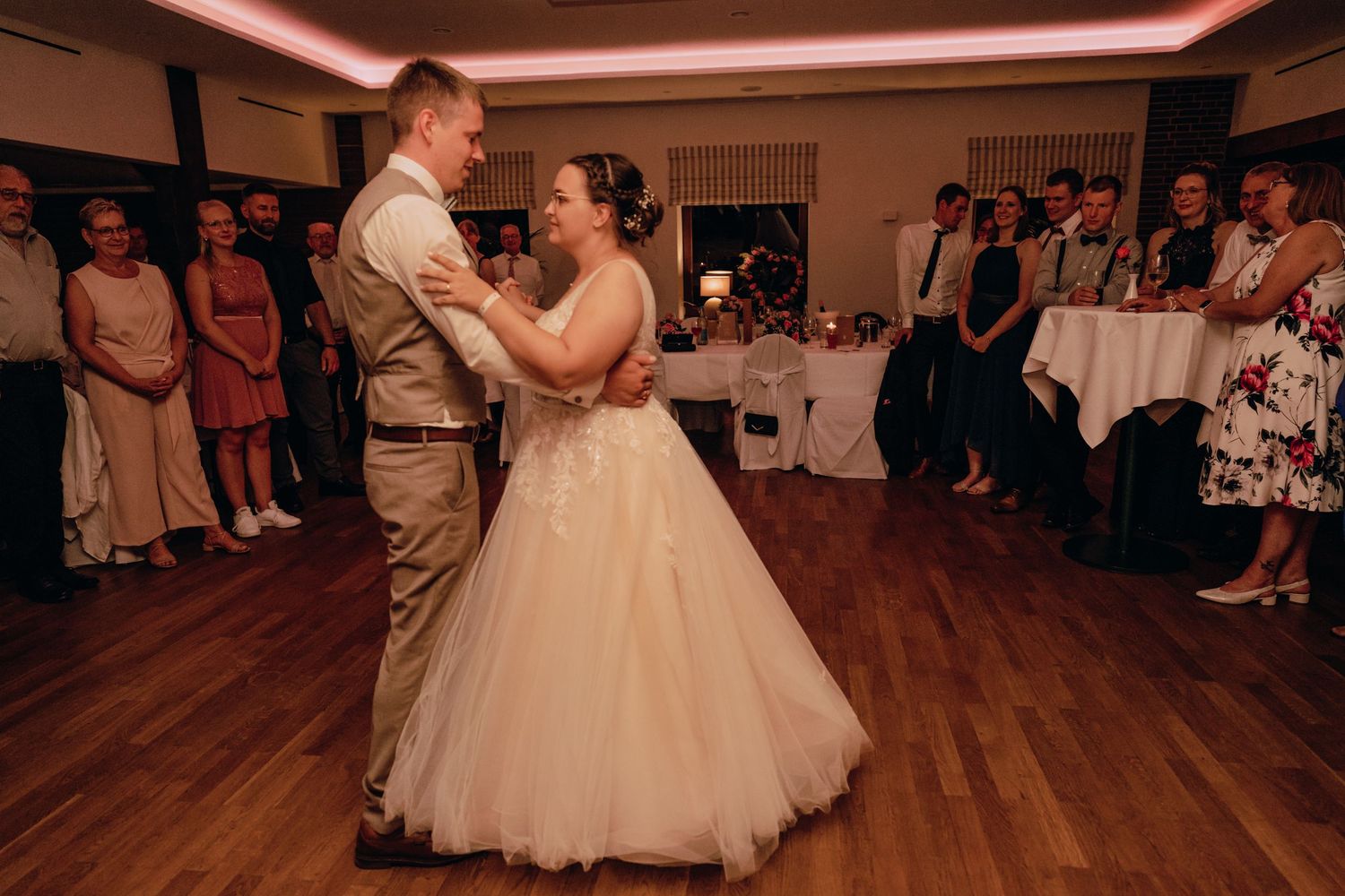 Newlyweds share their first dance on a wooden floor surrounded by guests at their wedding reception.