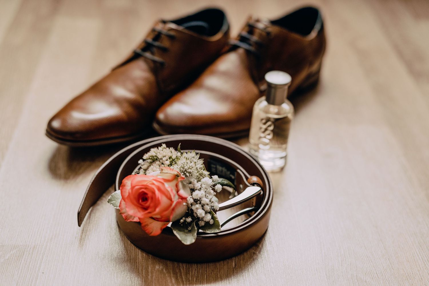 Brown leather dress shoes and floral wedding rings displayed on wooden surface for wedding detail photography.