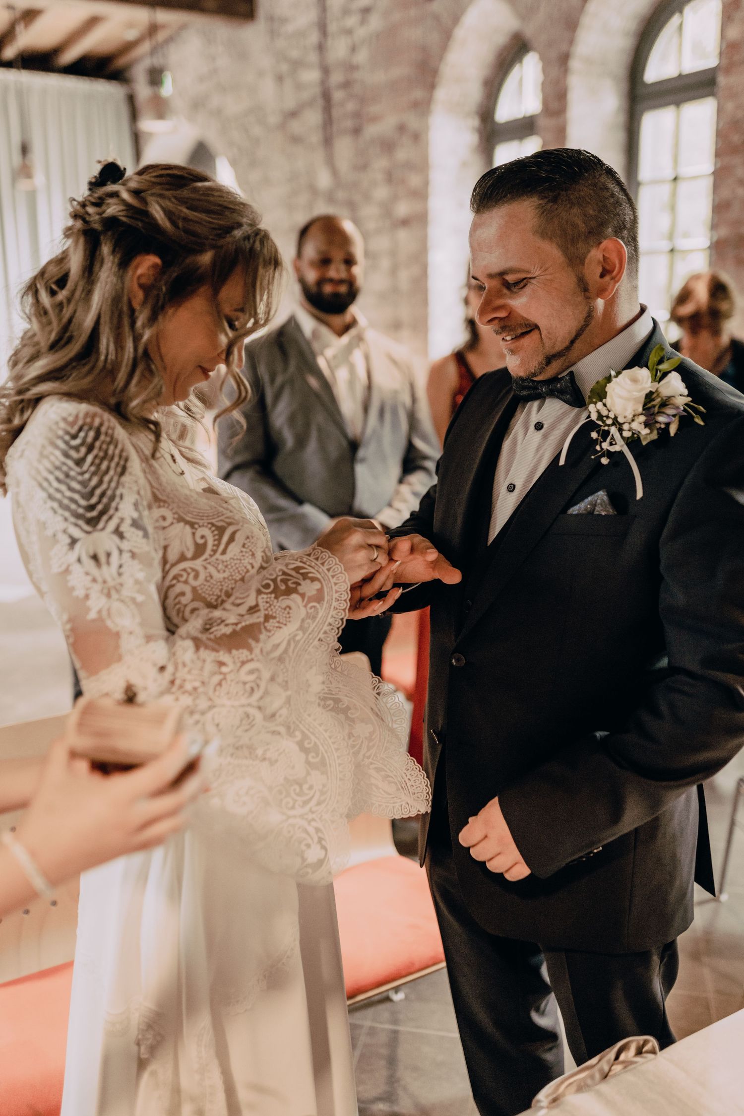 Two figures share an intimate moment during a wedding ceremony as they exchange rings in a beautifully lit indoor venue.
