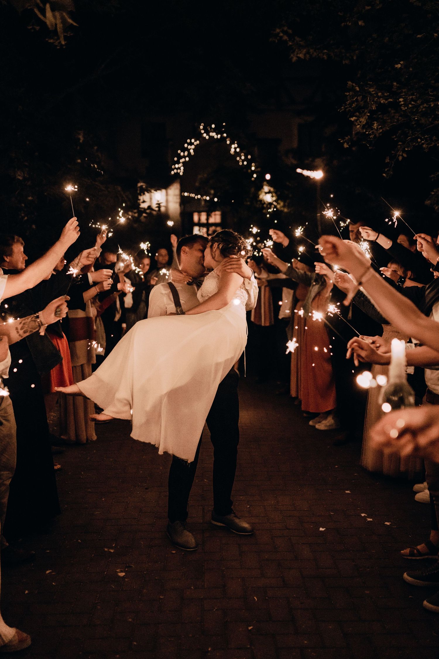 A romantic sparkler exit with a groom lifting his bride while guests create a tunnel of light at a nighttime wedding celebration.