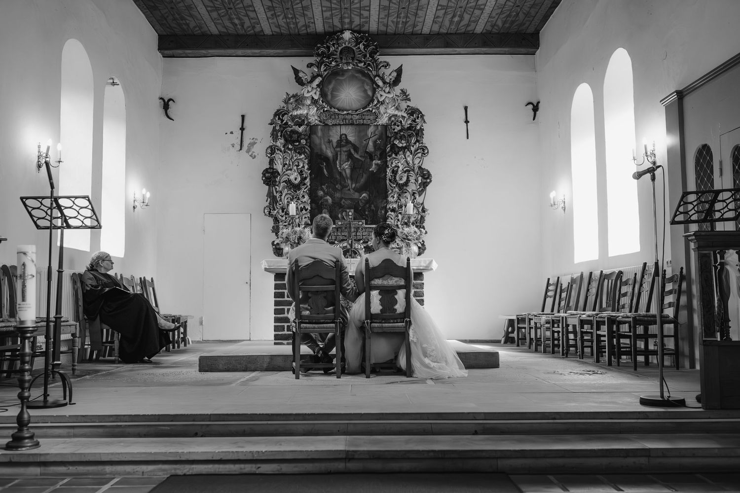 Black and white interior of a church showing ornate altar decorations, wooden chairs, and arched windows along white walls.