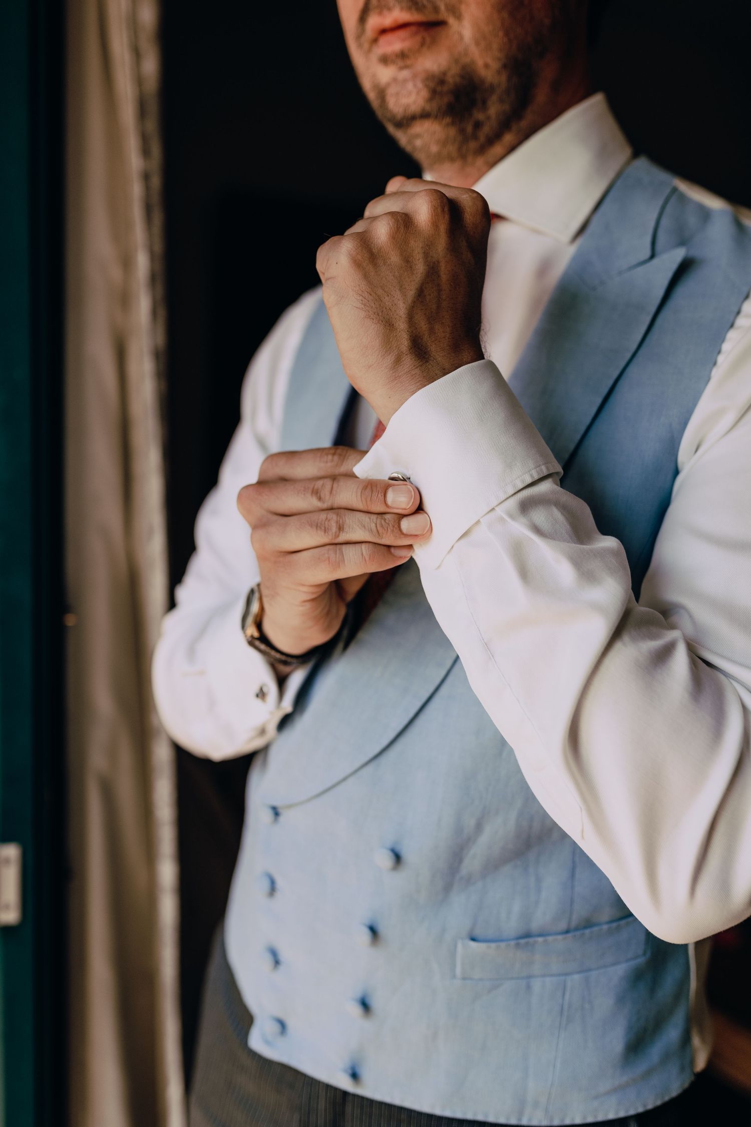 Close-up of groom adjusting formal light blue vest and white dress shirt cuffs before wedding ceremony.