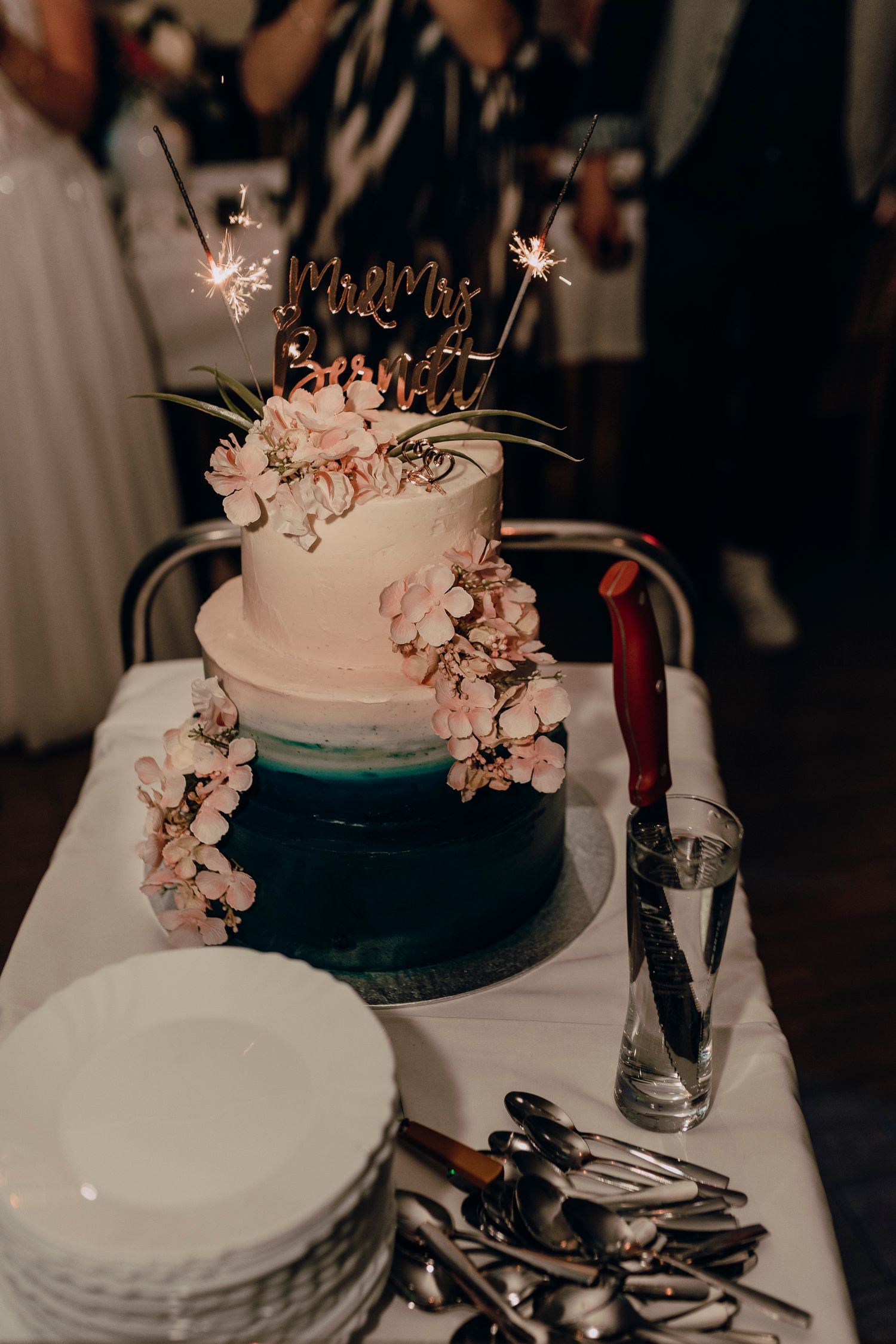 A wedding cake with teal and white tiers decorated with delicate pink cherry blossoms and sparkler toppers on display.