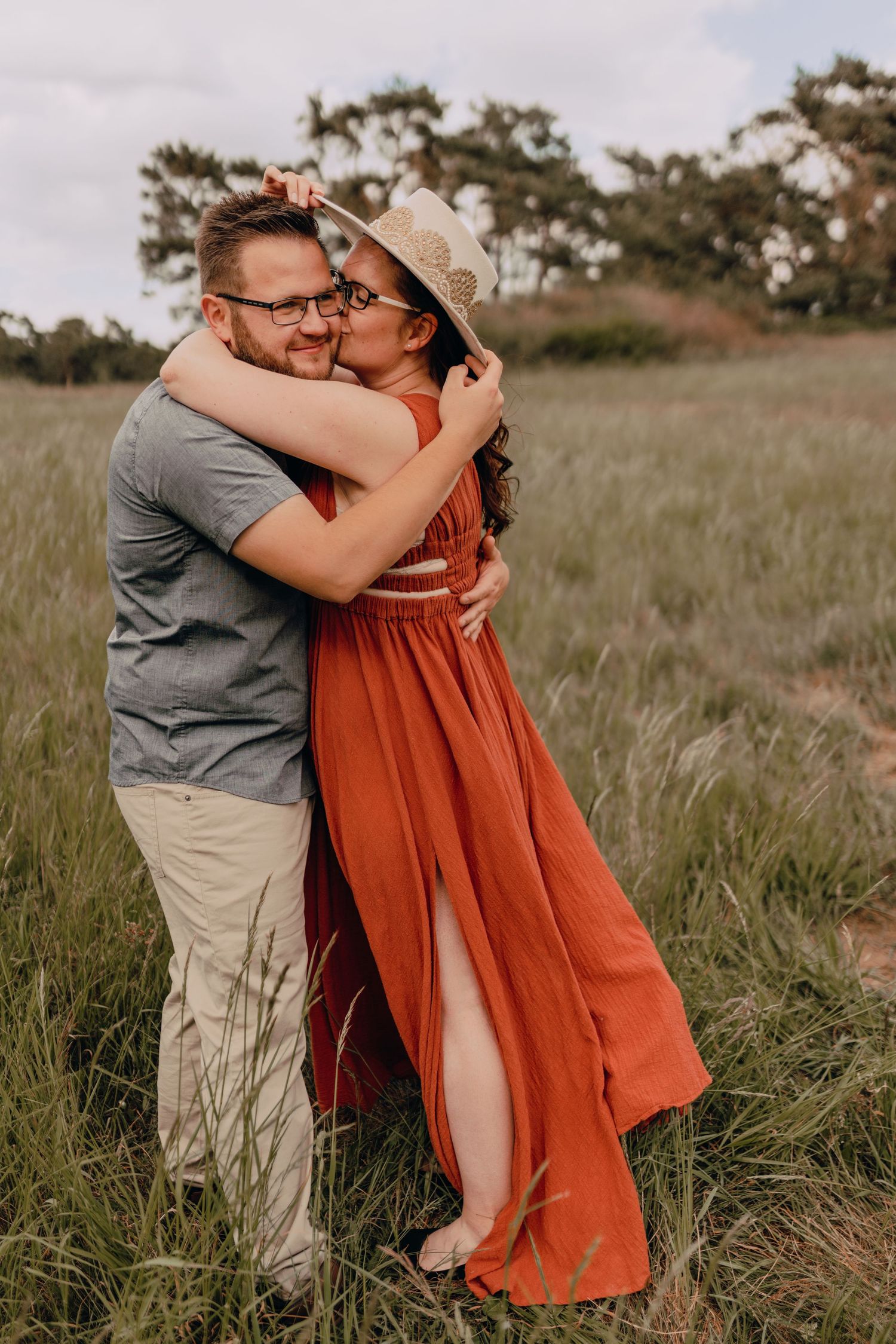 Romantic couple embraces in a sunlit meadow with tall grass, her orange dress flowing in the breeze.