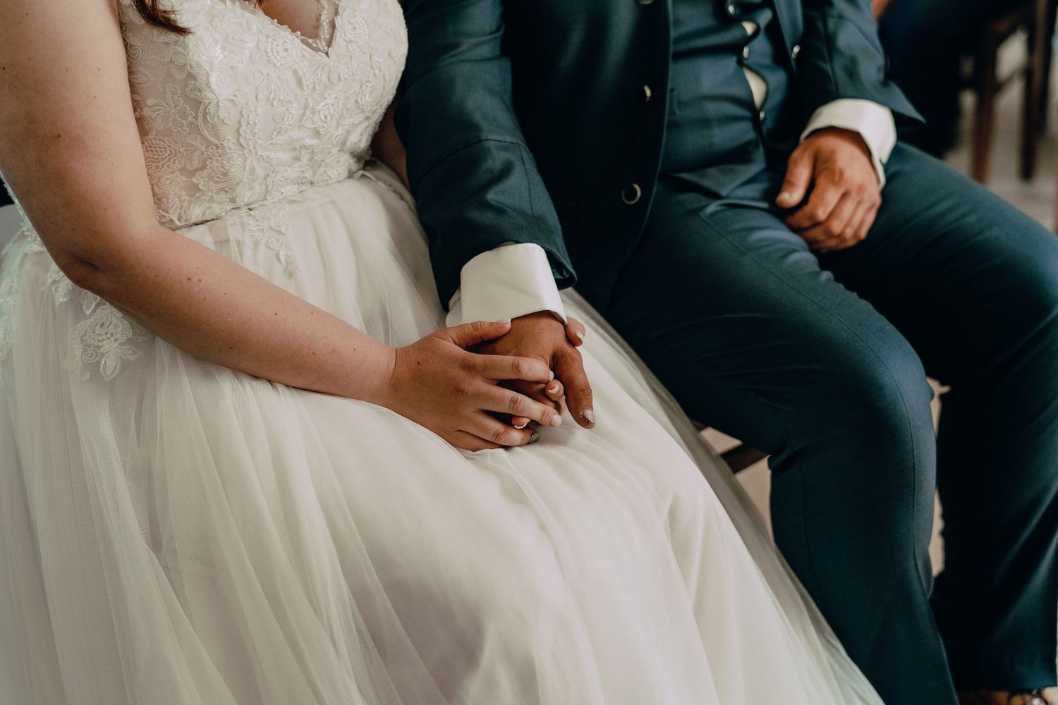 Close-up view of a couple holding hands at their wedding, showing white dress and dark suit details.