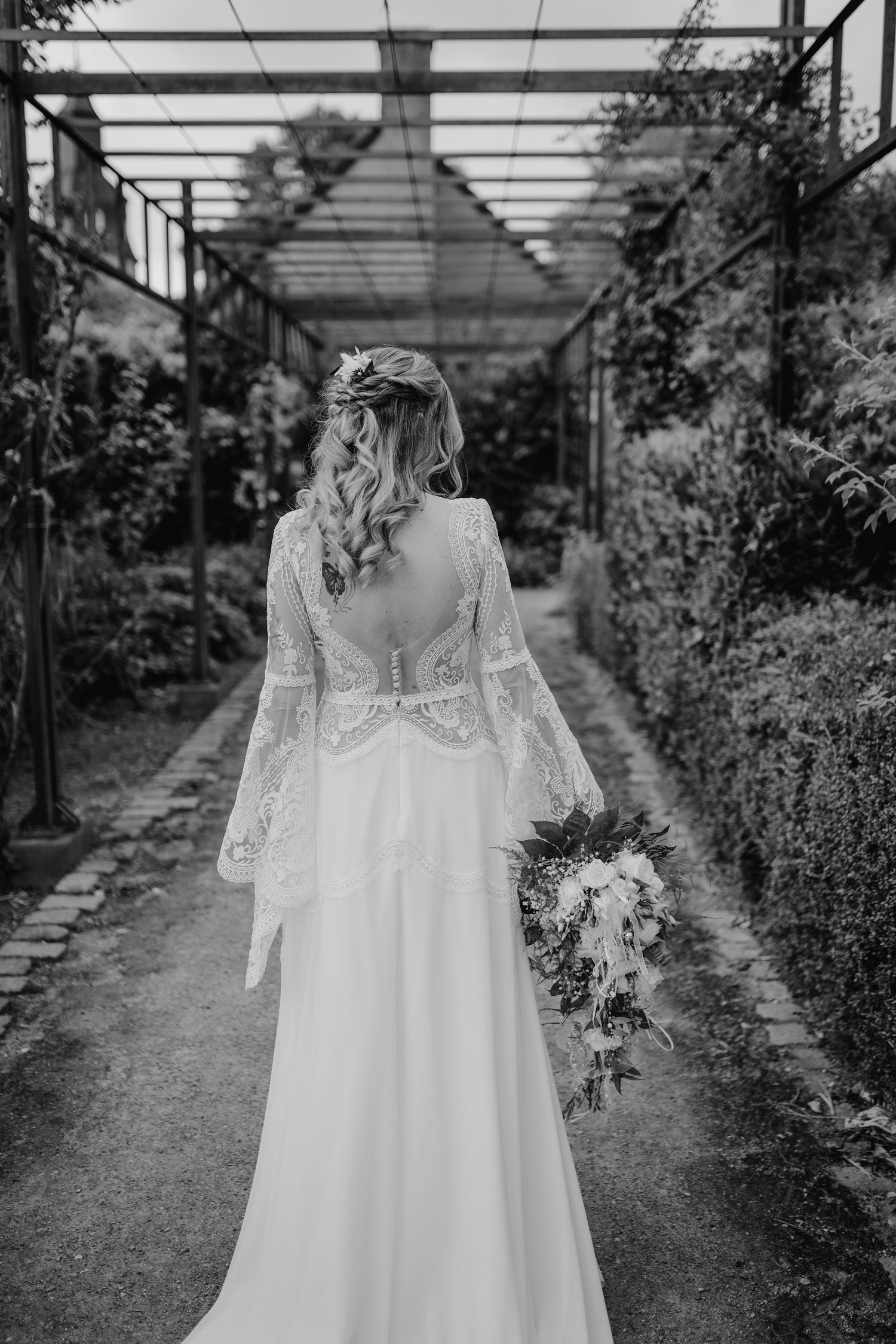 Black and white photo of bride in lace back wedding gown with flowing sleeves holding bouquet in garden pathway.
