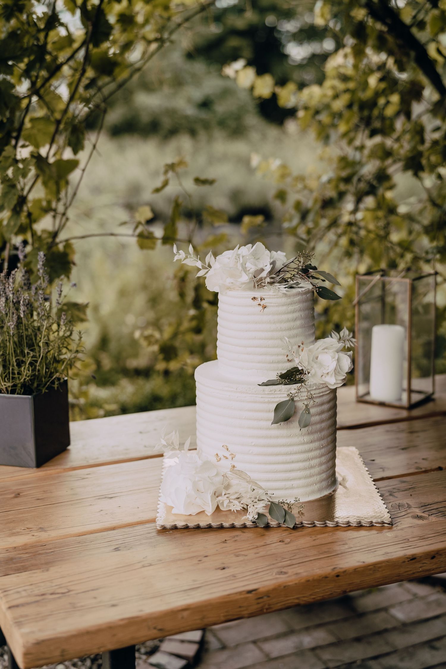Two-tiered white textured wedding cake decorated with fresh flowers on wooden table outdoors.