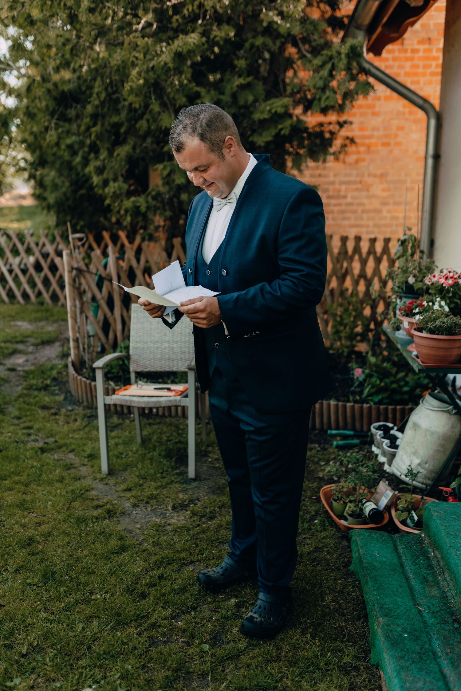 Person in a navy suit reads a document while standing in a garden with lattice fencing and potted plants.