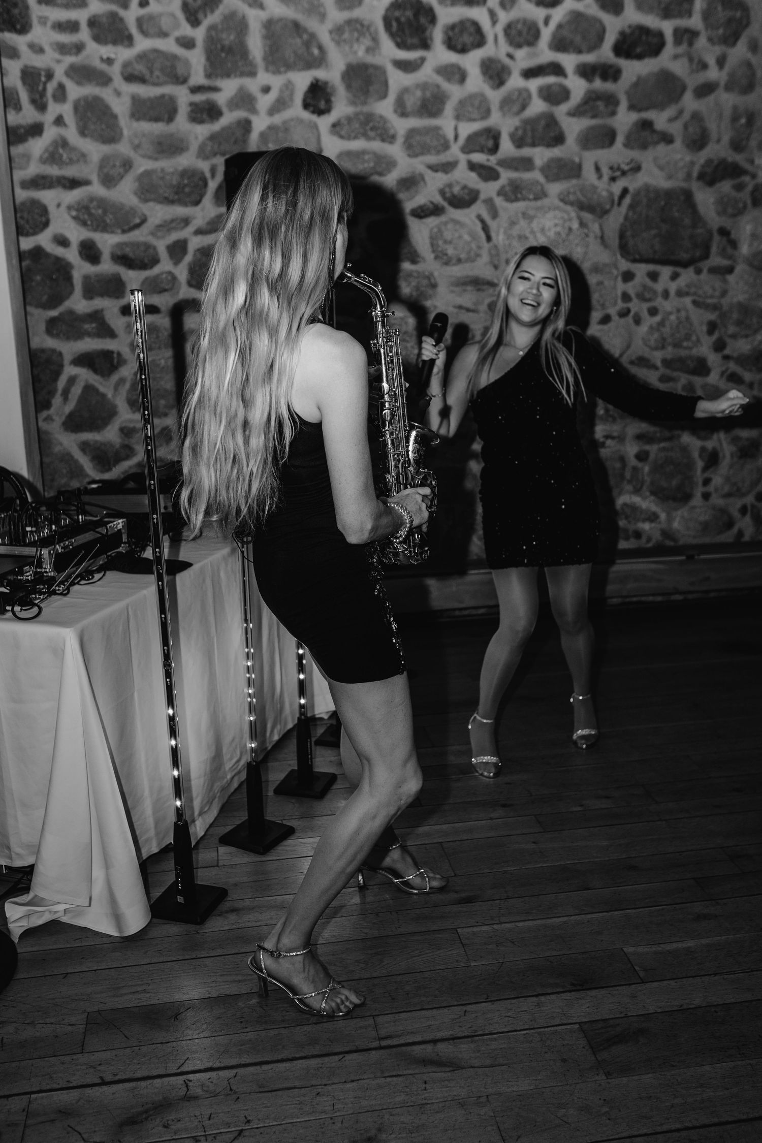 Black and white photo showing women dancing and socializing at a party with a stone wall backdrop.