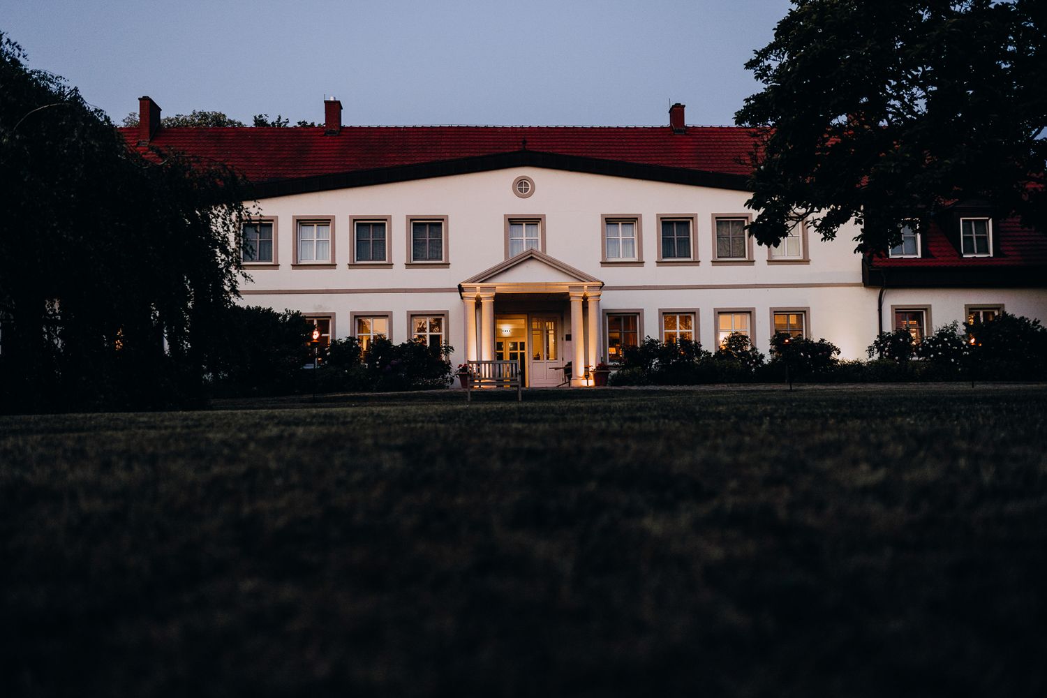 Large white manor house with pillared entrance illuminated at dusk.