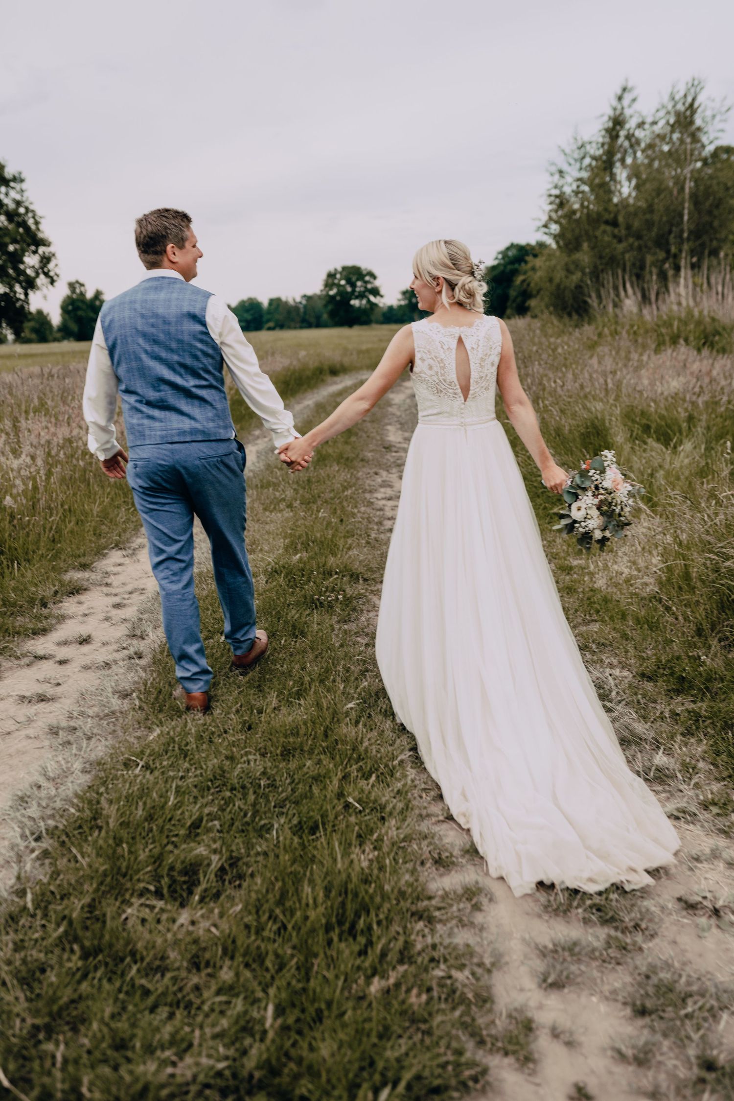 Bride and groom in wedding attire walk up sandy path in rustic outdoor setting with wildflowers and natural landscape.