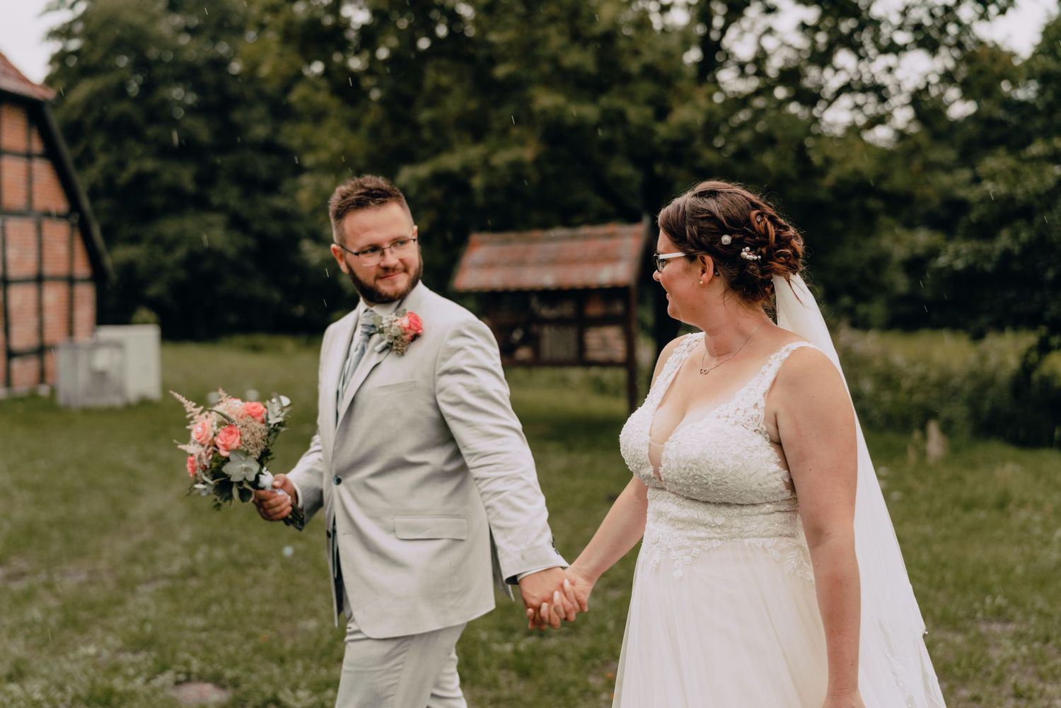 Newlyweds walking hand in hand outdoors with rustic barn and trees in the background.