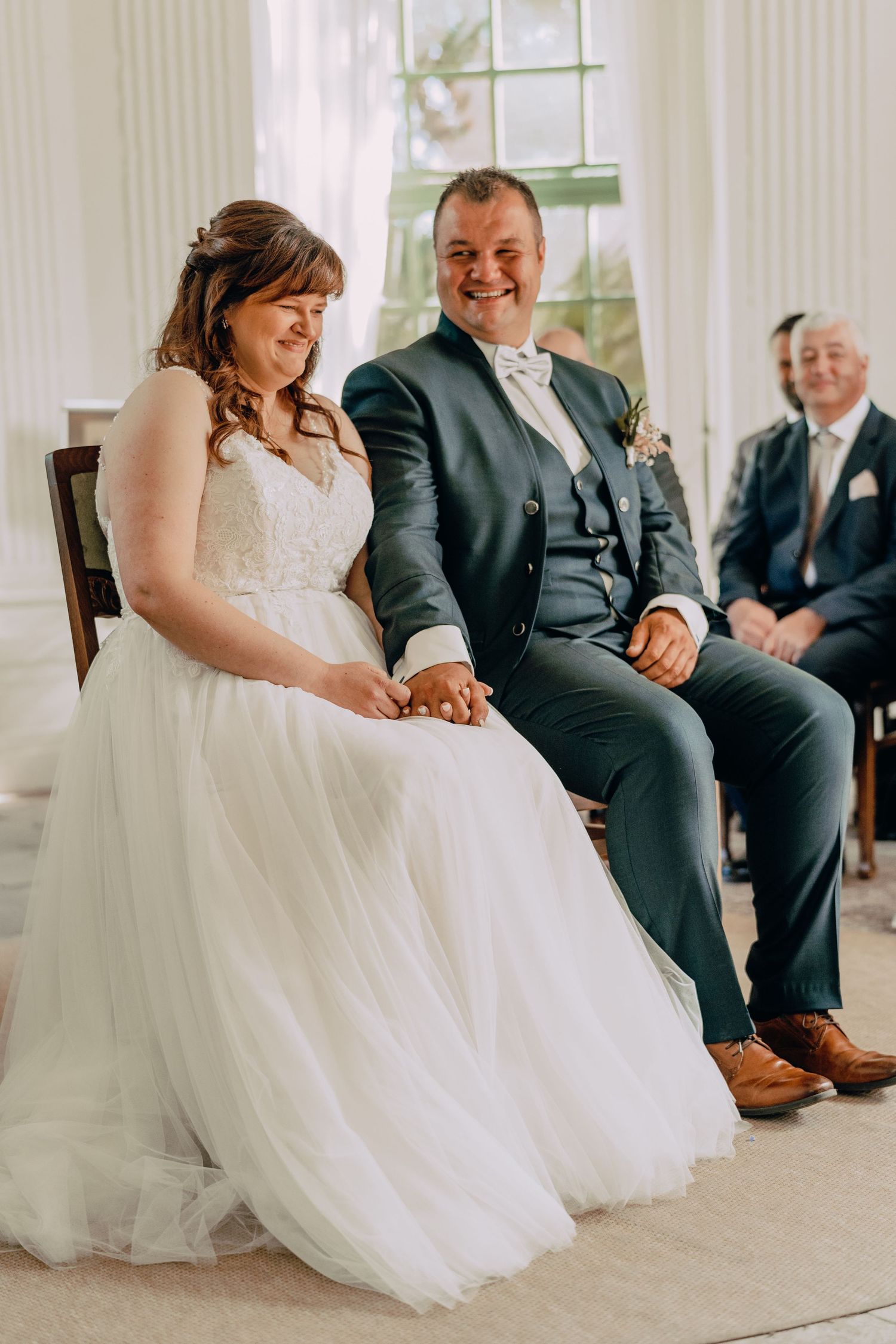 A couple sits together during their wedding ceremony while holding hands with a suited guest in the background.