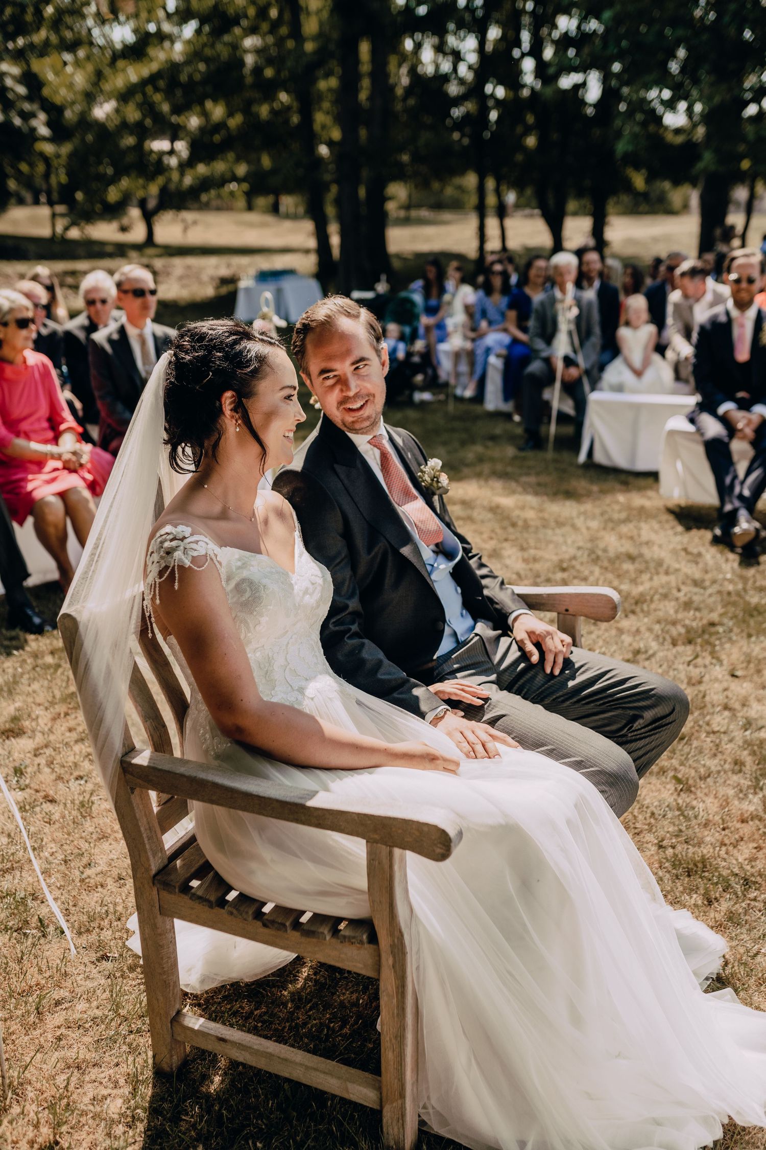 Couple seated during outdoor wedding ceremony with guests visible in background.