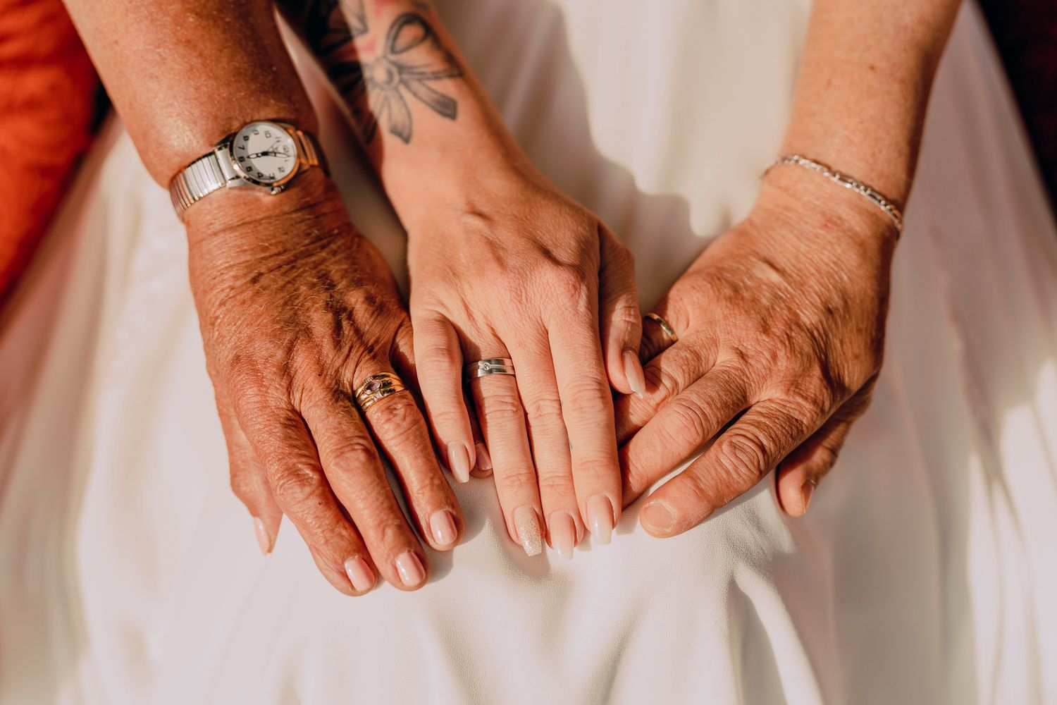 Three pairs of hands gently resting on white fabric with visible rings, tattoos, and jewelry.