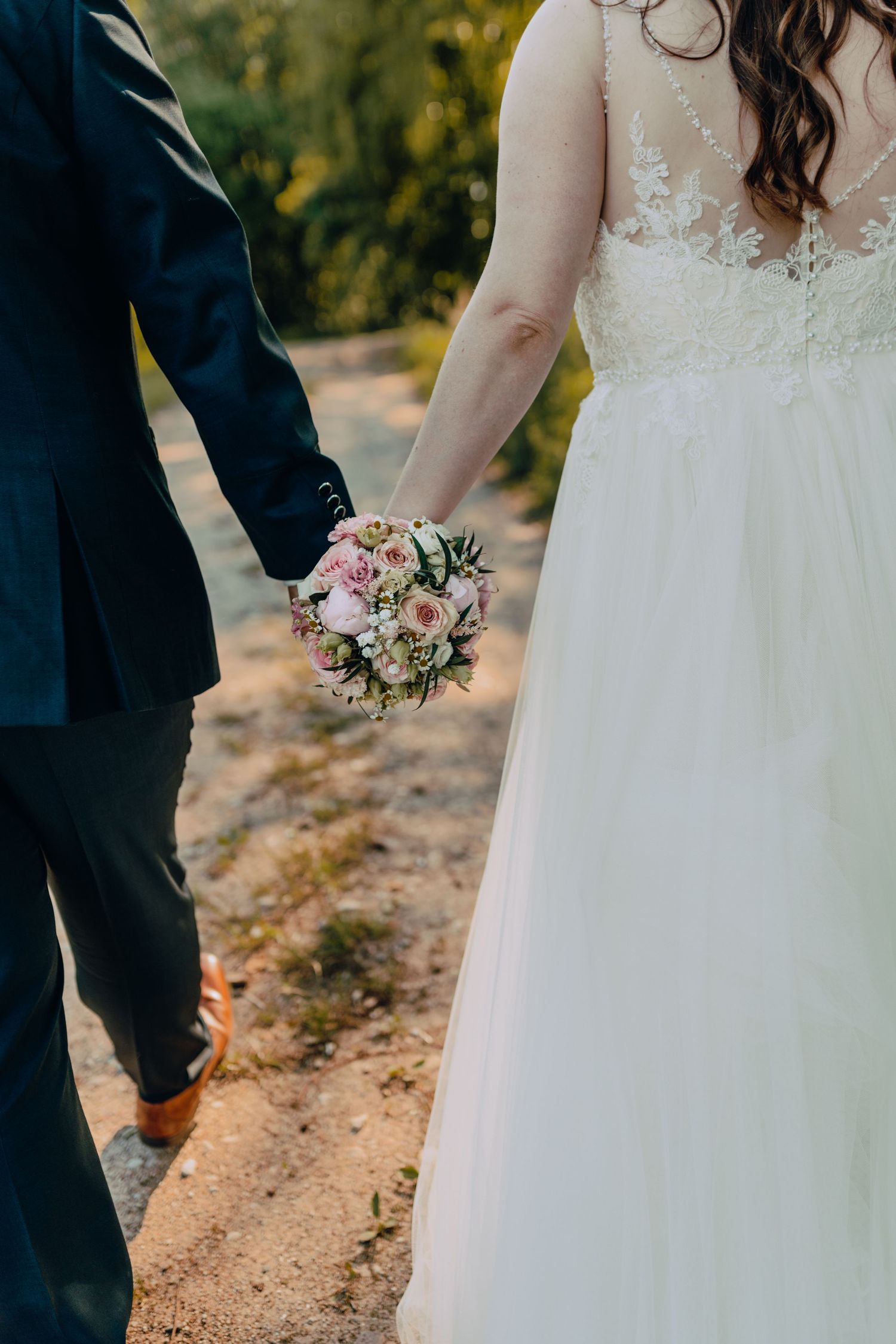 A close-up of a wedding couple holding hands with a pink and white flower bouquet between them.