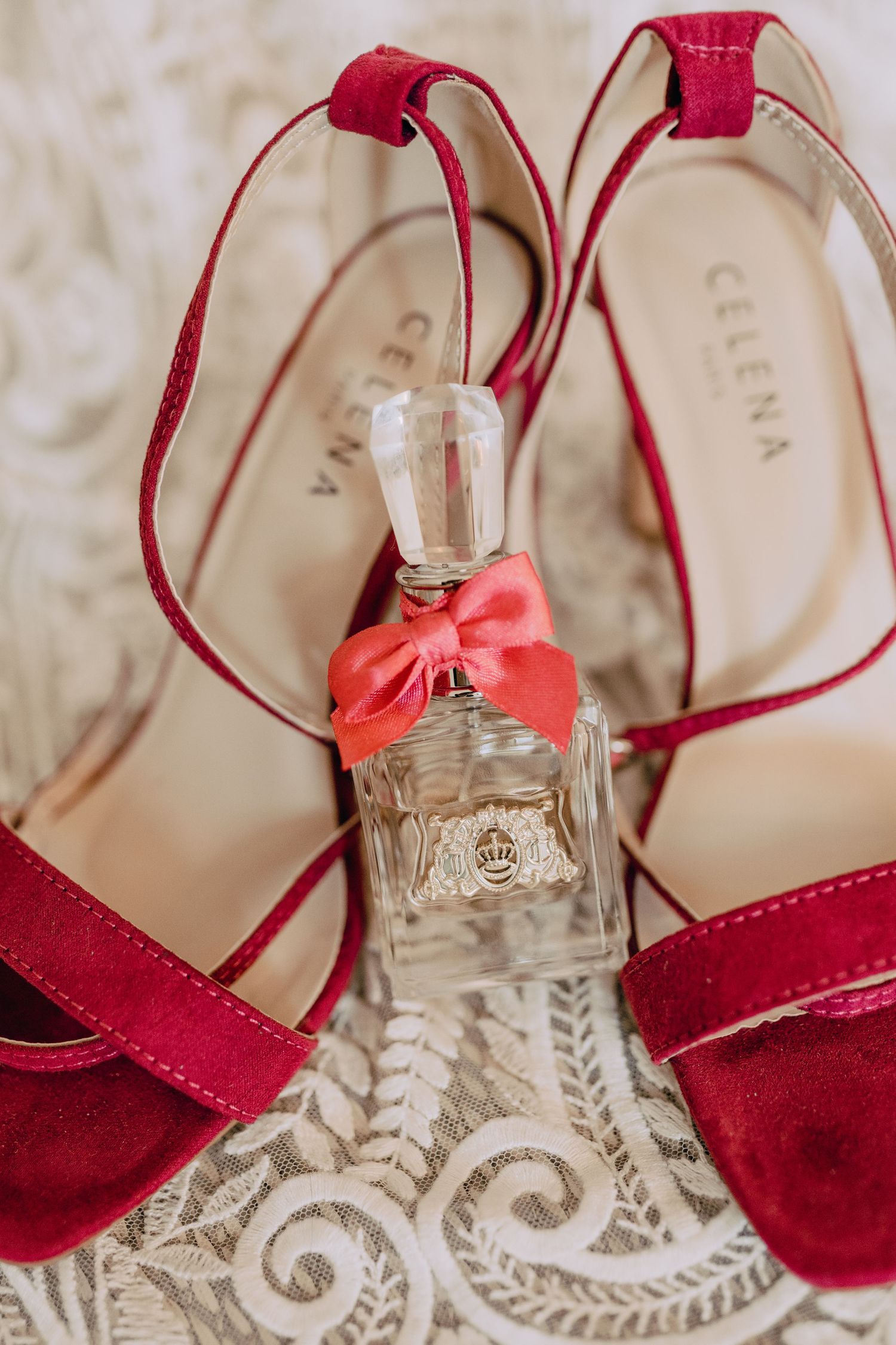 Red velvet high heel wedding shoes arranged with a perfume bottle adorned with a coral bow on lace background.