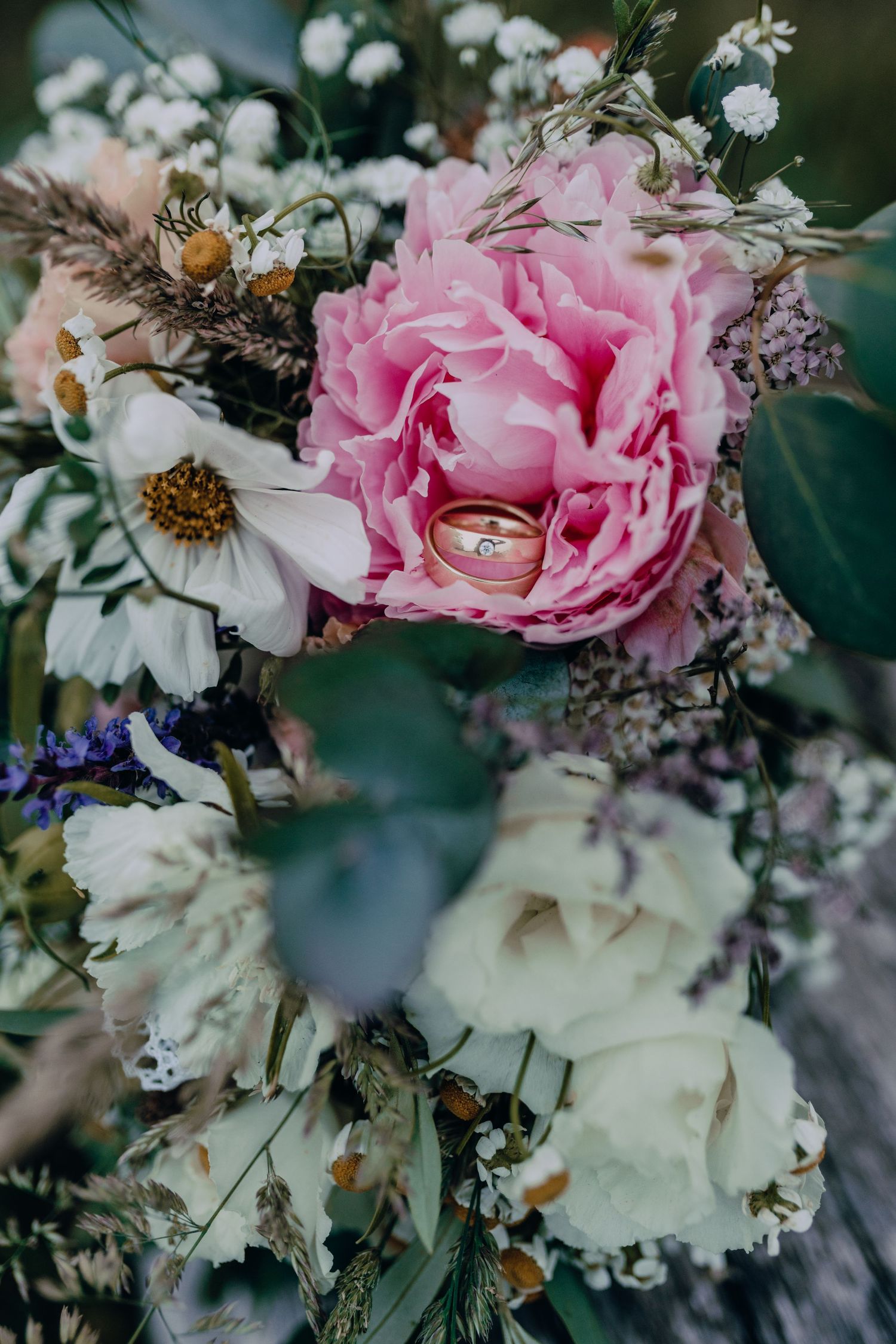 Wedding rings nestled in a lush floral arrangement of pink peonies, white roses, and delicate purple flowers with greenery.