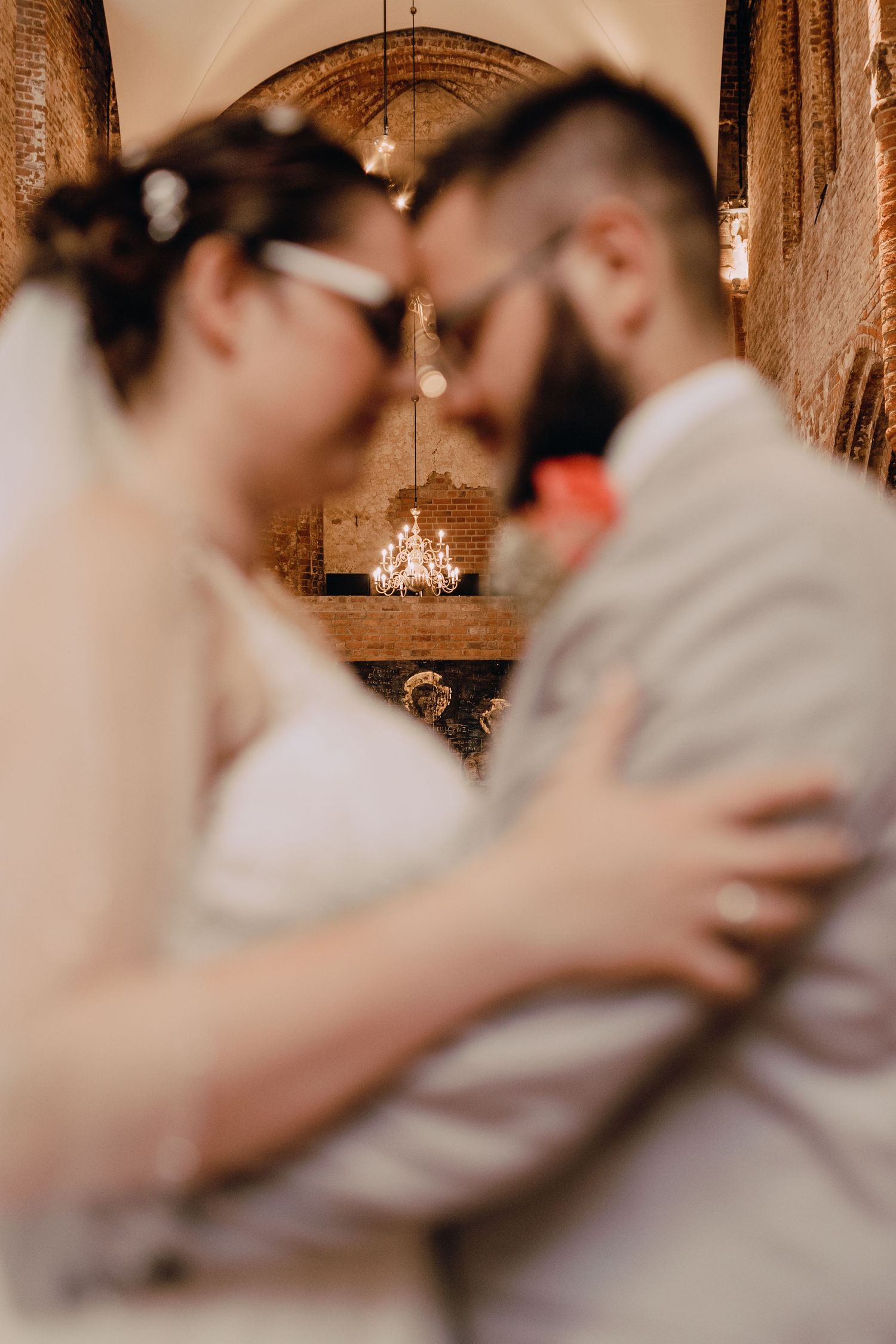 Couple embracing in a rustic venue with elegant chandeliers and romantic lighting during their wedding celebration.