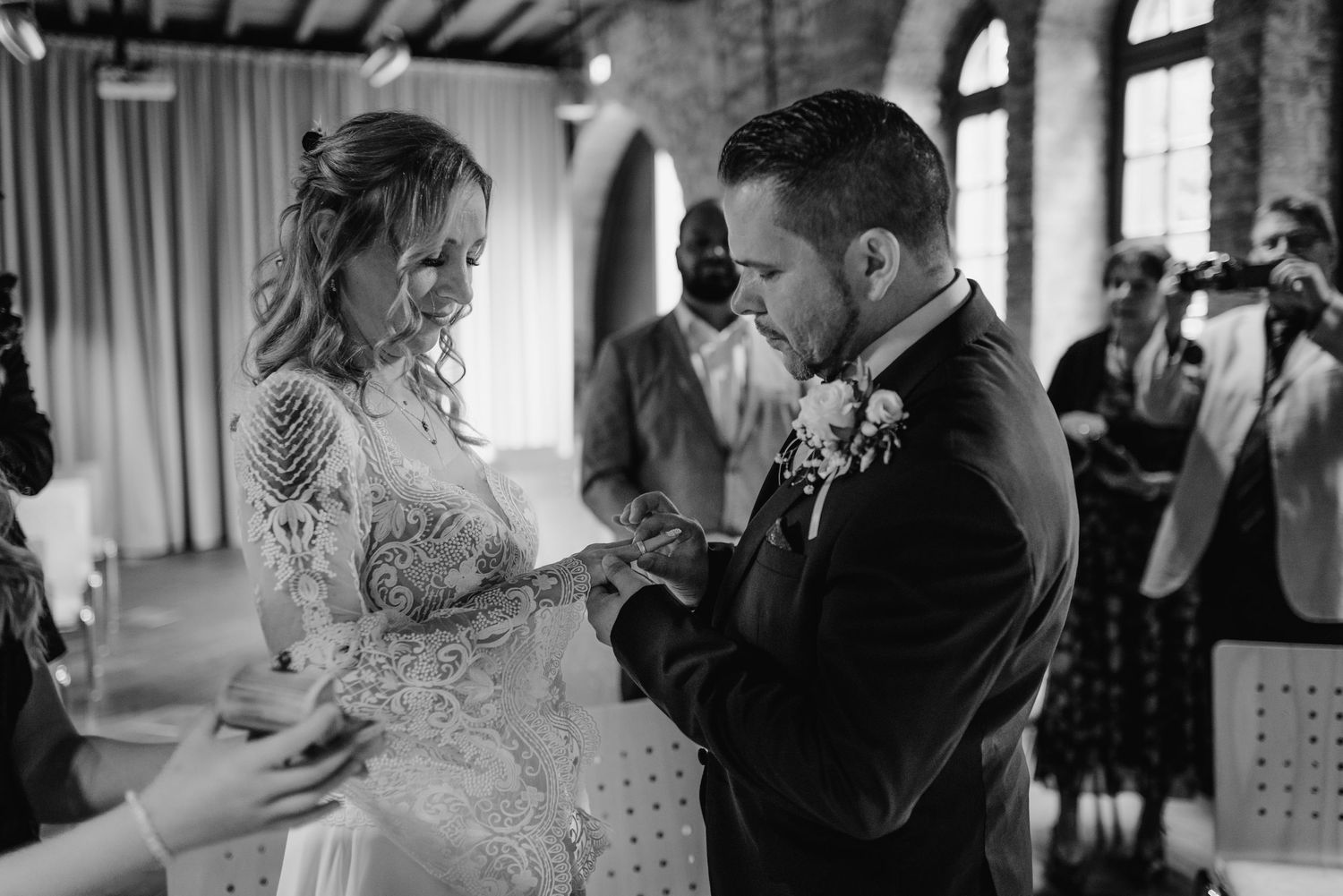 Black and white photo of a wedding ceremony showing bride in lace gown and groom exchanging rings during the vows.