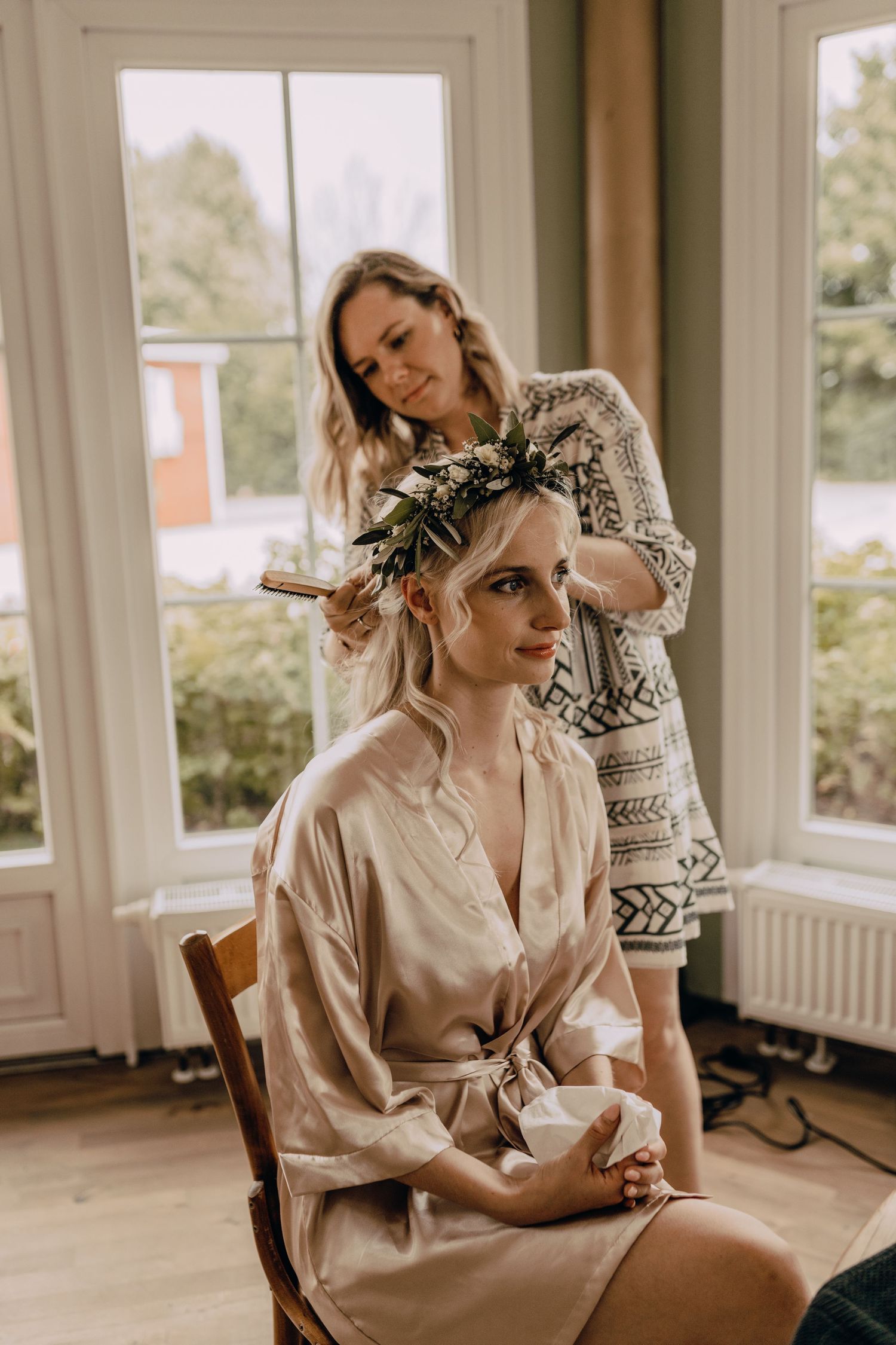 Wedding hair and makeup preparation in a sun-lit room with greenery crown details.