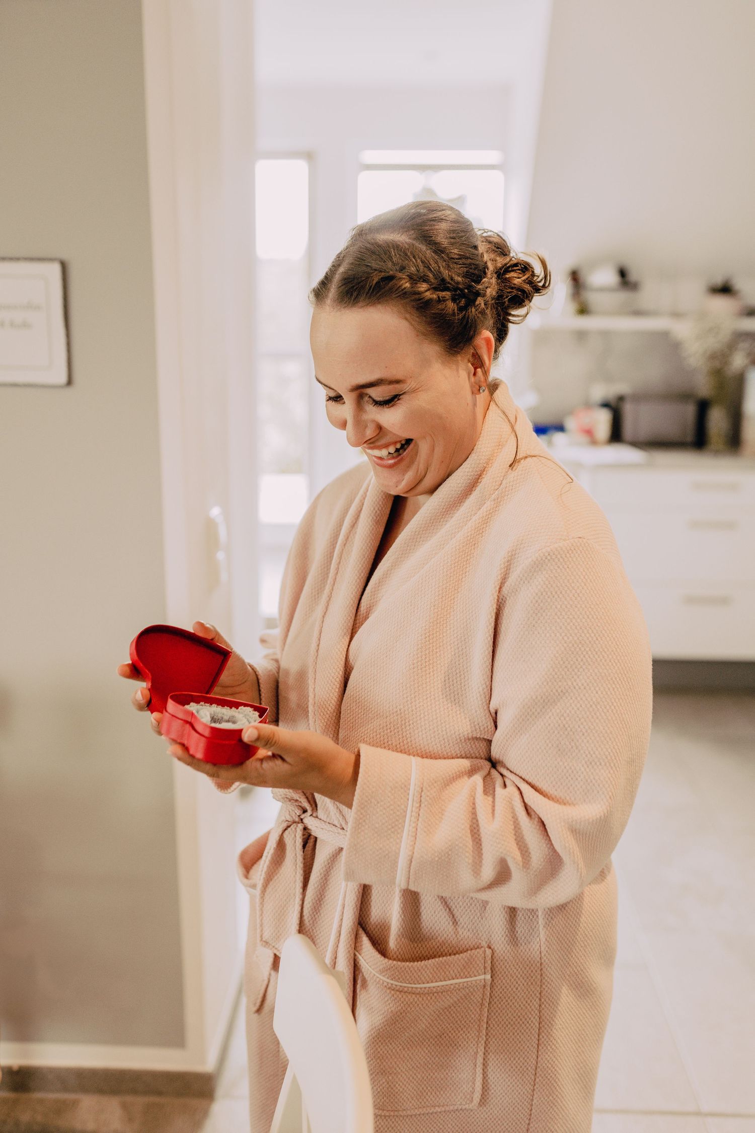 A person in a peach-colored robe smiles while opening a small red gift box in a bright interior space.