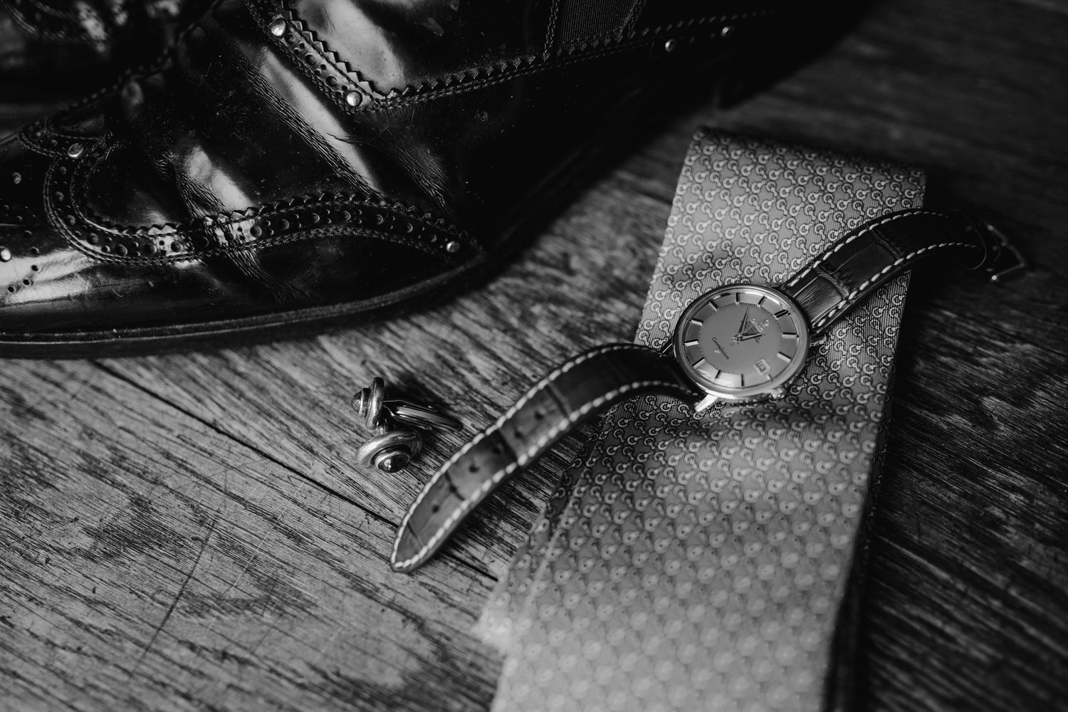 Vintage wristwatch and brogue shoe detail on wooden surface in black and white.