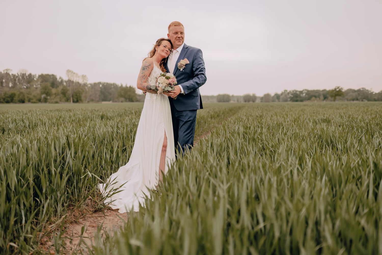 A romantic couple embraces on a path through a lush green wheat field on their wedding day.