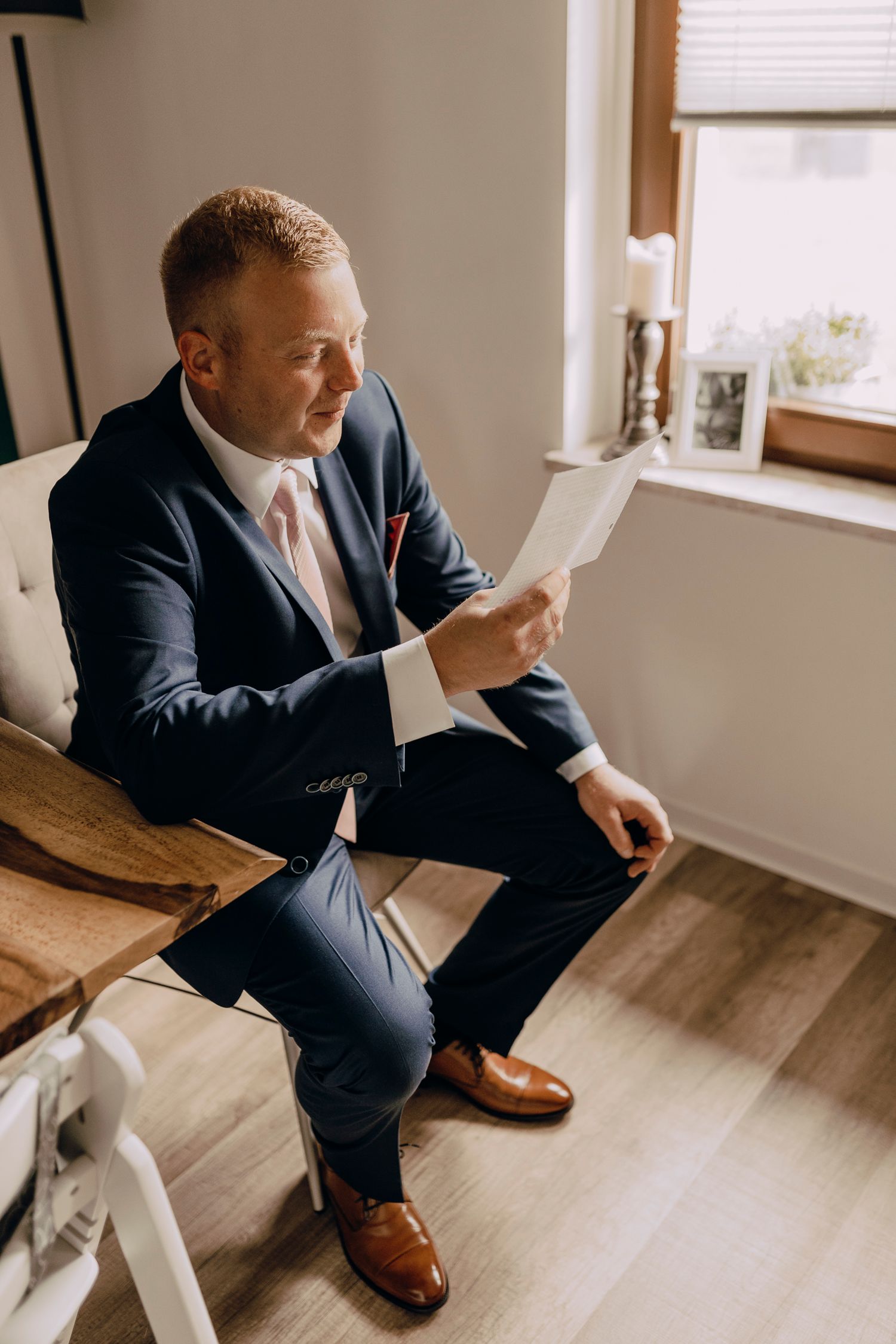 Someone in a navy suit and brown leather shoes checks their phone while sitting in a bright room near a window.