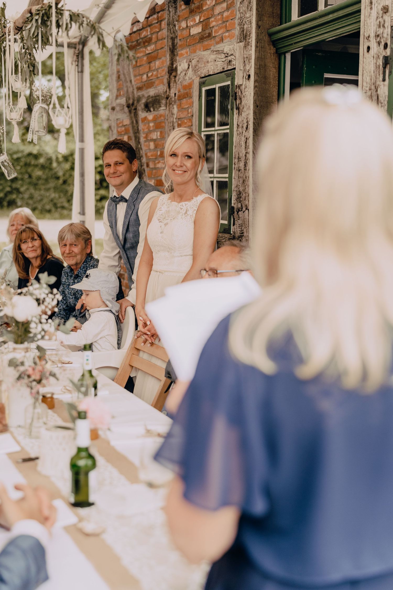 Wedding reception dining setup with long tables under hanging greenery at a rustic brick venue.
