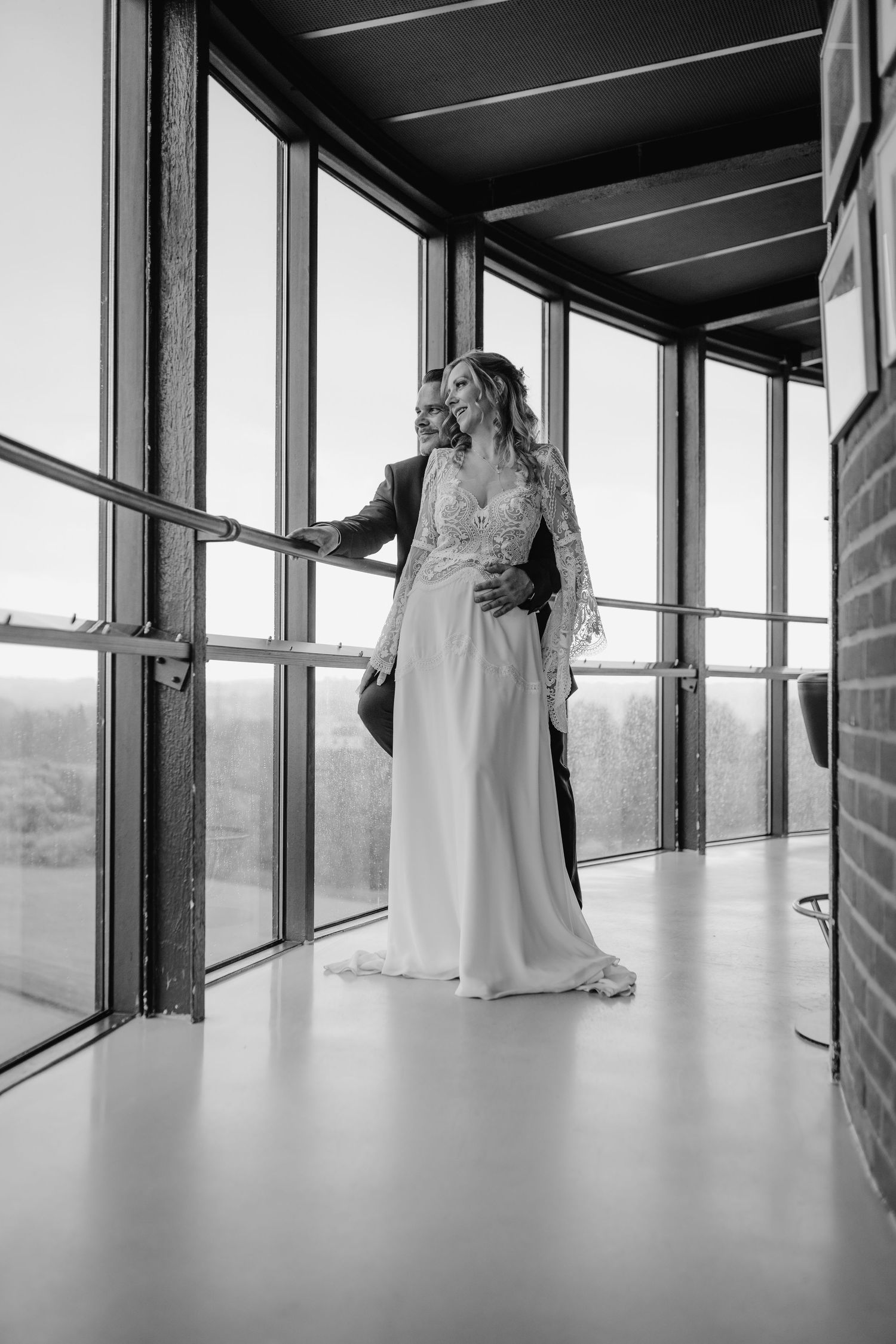 A person in a flowing white wedding gown stands by large windows in a modern architectural space, photographed in black and white.