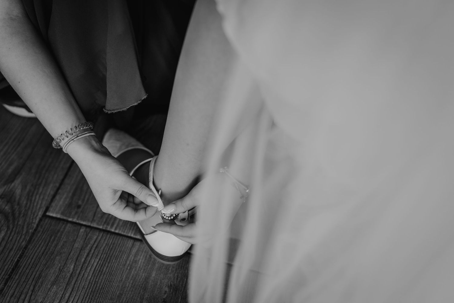 A person adjusts white ballet flats on their feet while wearing a flowing dress against a dark wooden floor.