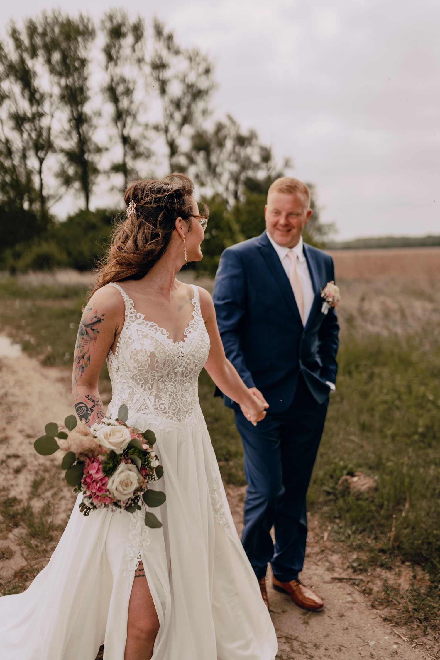 A couple strolls hand in hand on a sandy path through a grassy meadow during wedding photos.