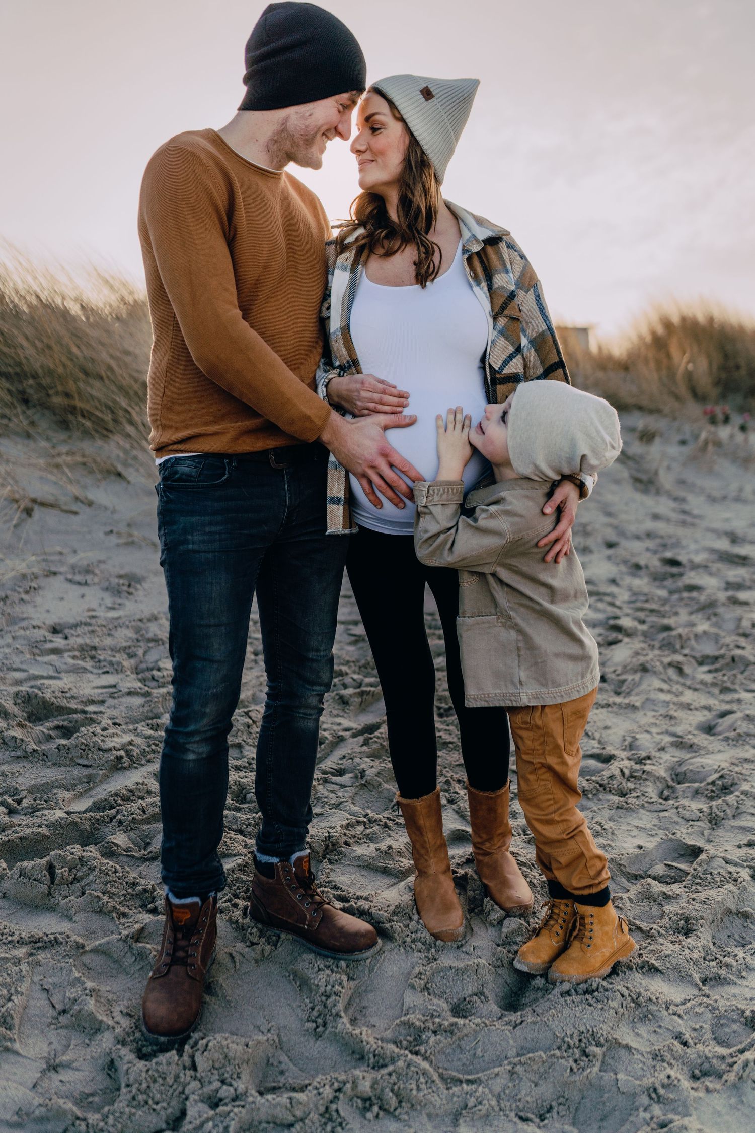 Expecting parents embrace on a sandy beach while a young child in brown pants looks on.
