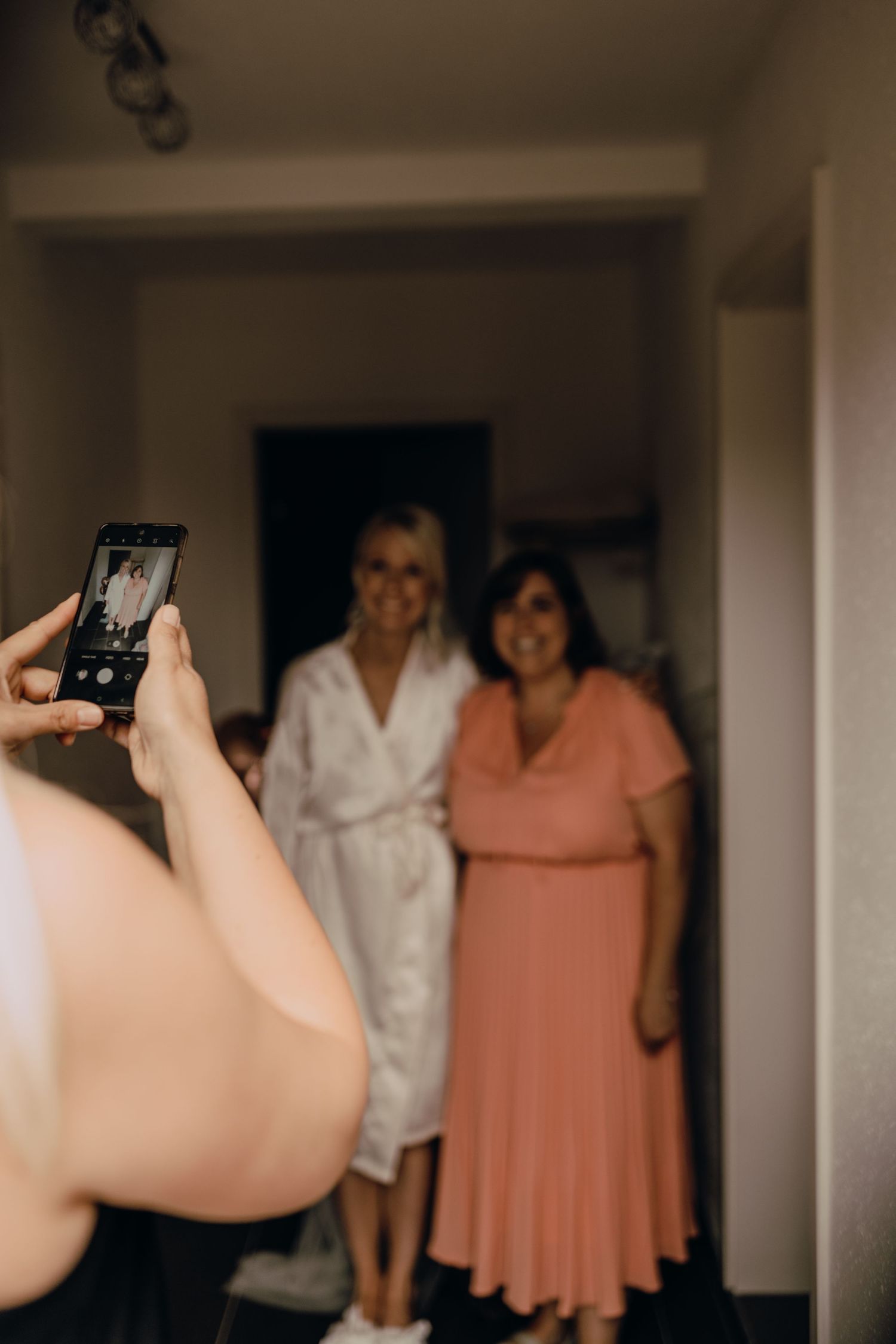 Someone taking a phone photo of two people posing together in a hallway.