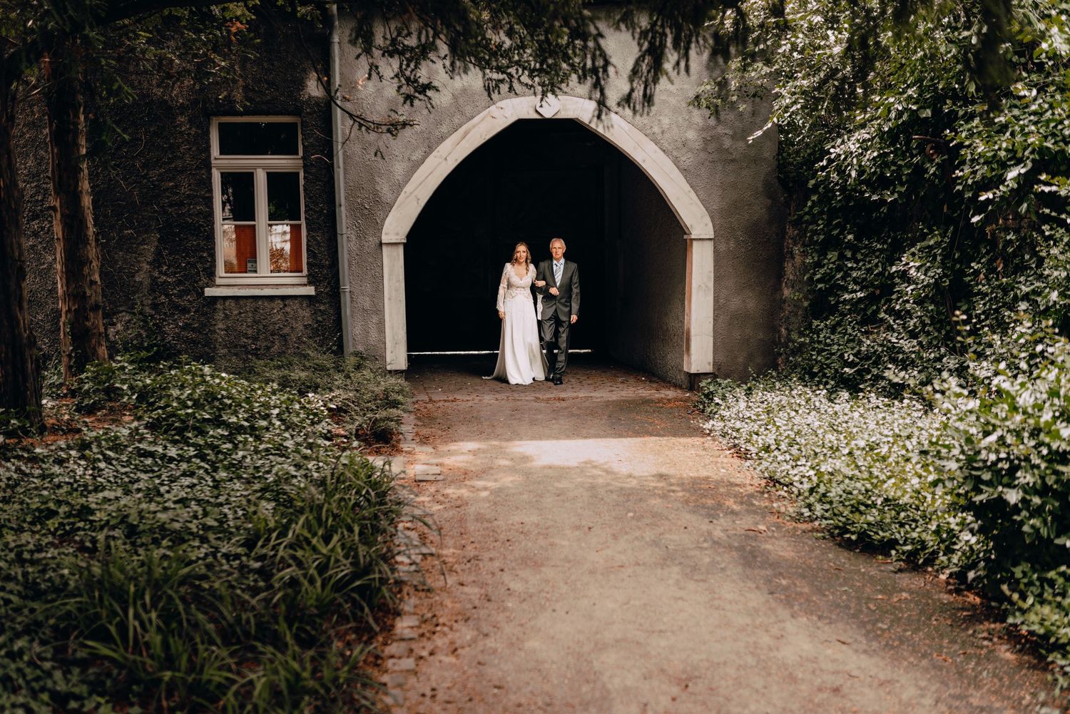 A couple stands under a stone archway surrounded by lush greenery on a dirt path.