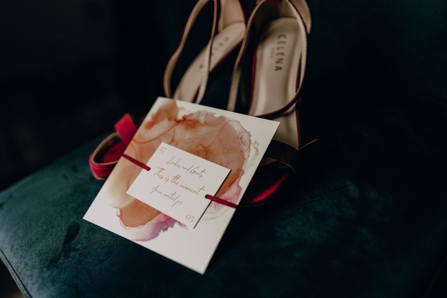 Wedding invitation and blush colored heels arranged on a dark velvet surface.