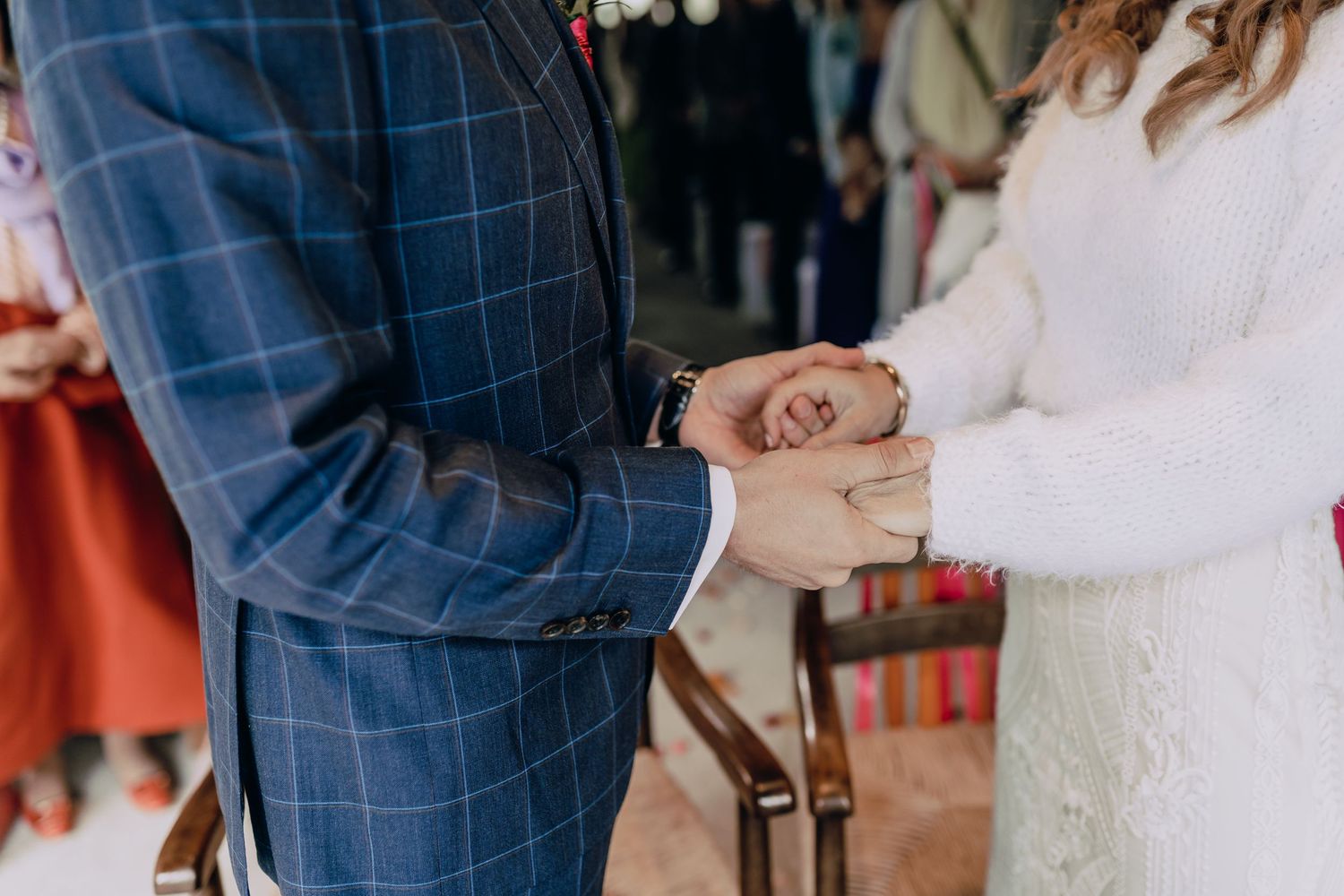 Close-up of couple's hands clasped together, with groom wearing blue plaid suit and bride in white lace.