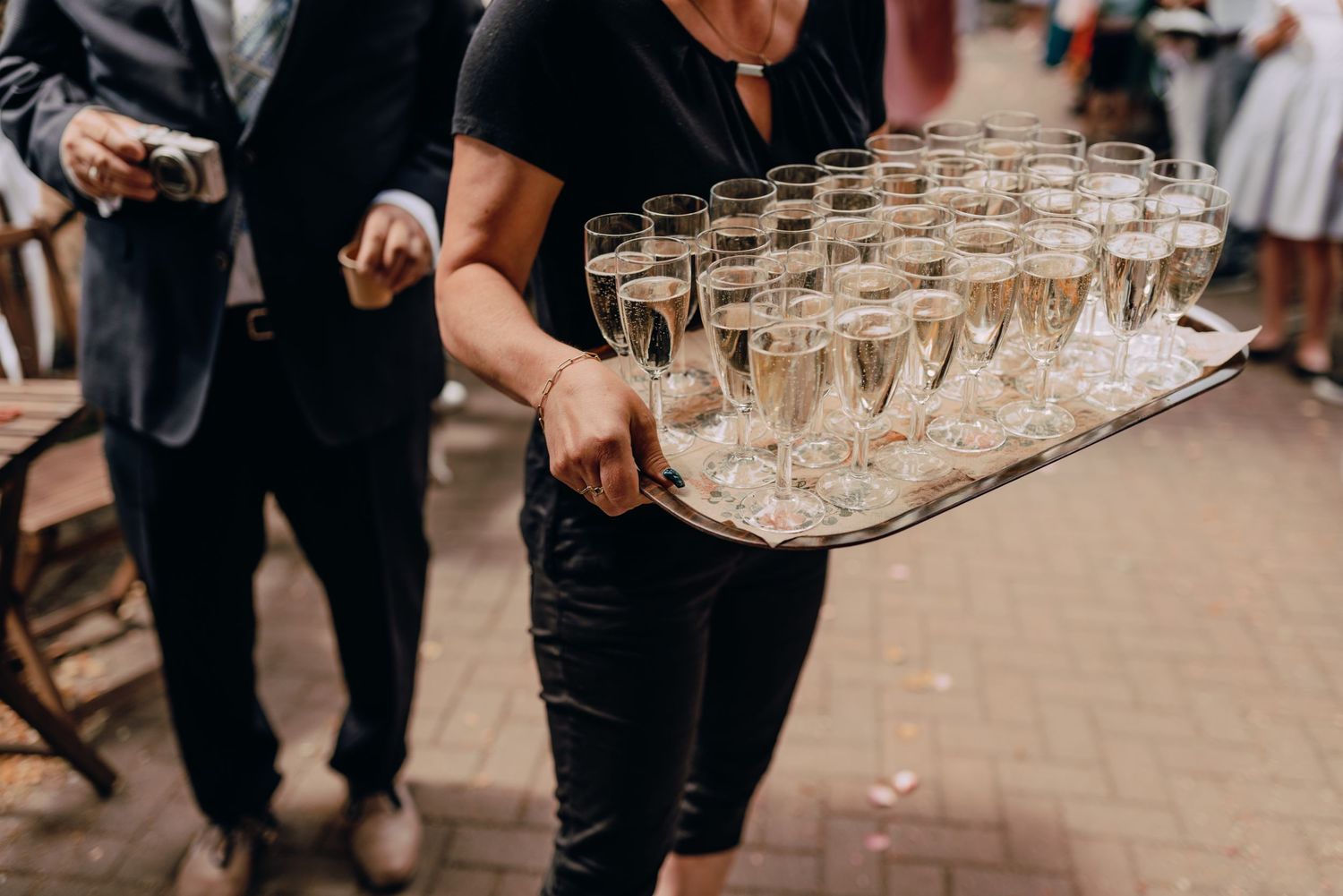 Server carries tray of champagne glasses at formal event through elegantly dressed crowd of guests.