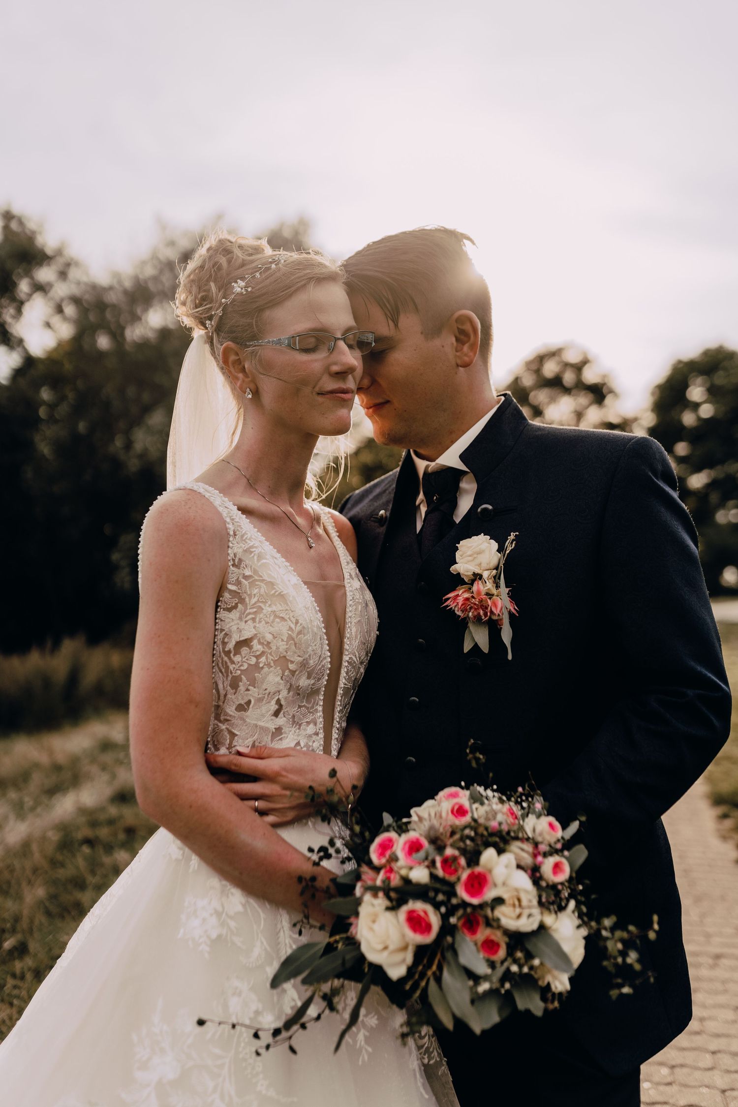 A romantic outdoor wedding portrait at sunset with a bouquet of pink roses and a classic black tuxedo and white gown.