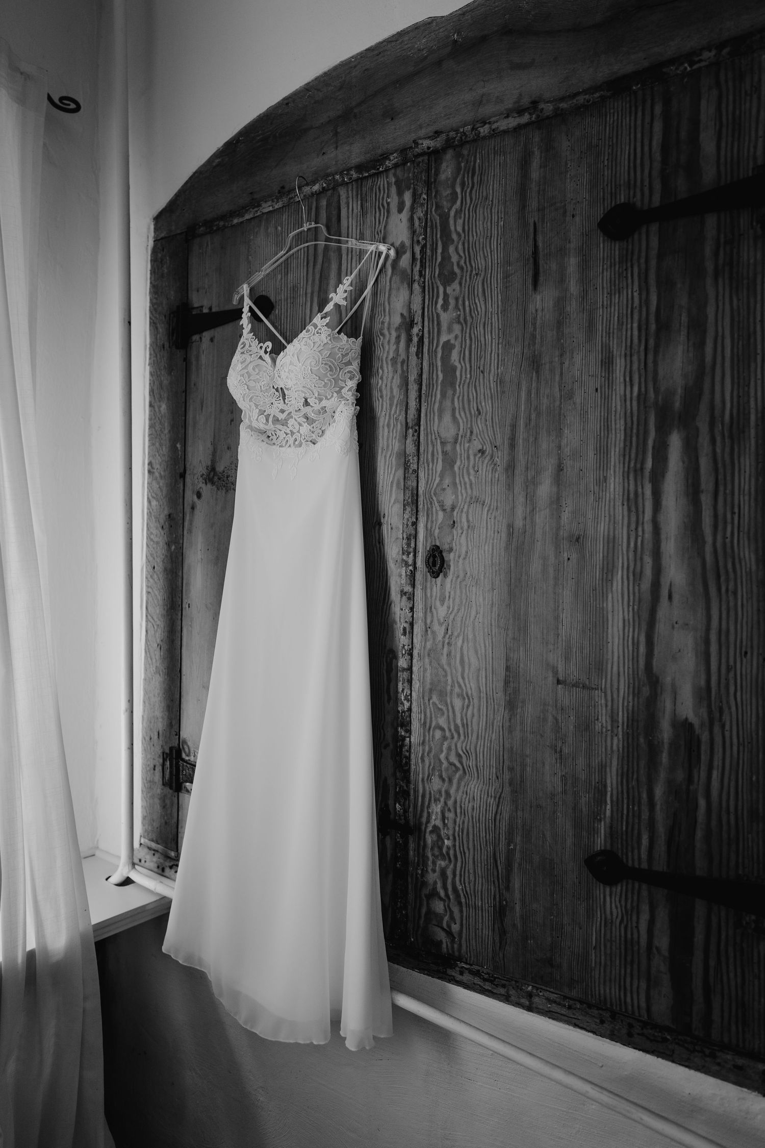 A white wedding dress with delicate lace bodice hangs elegantly against a rustic wooden door in black and white.