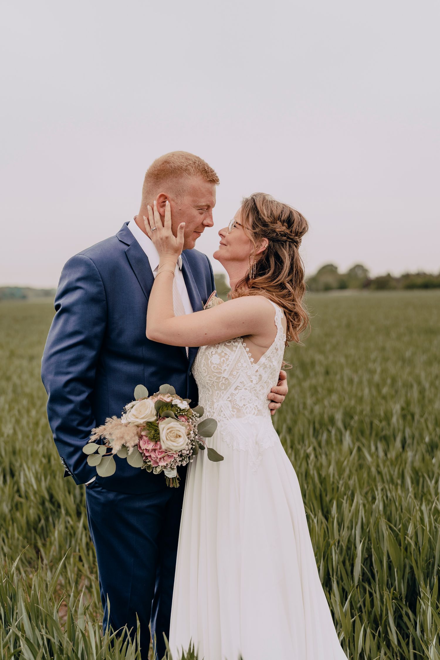 A romantic wedding photo in a green wheat field of a couple in navy suit and white dress holding a pink bouquet.
