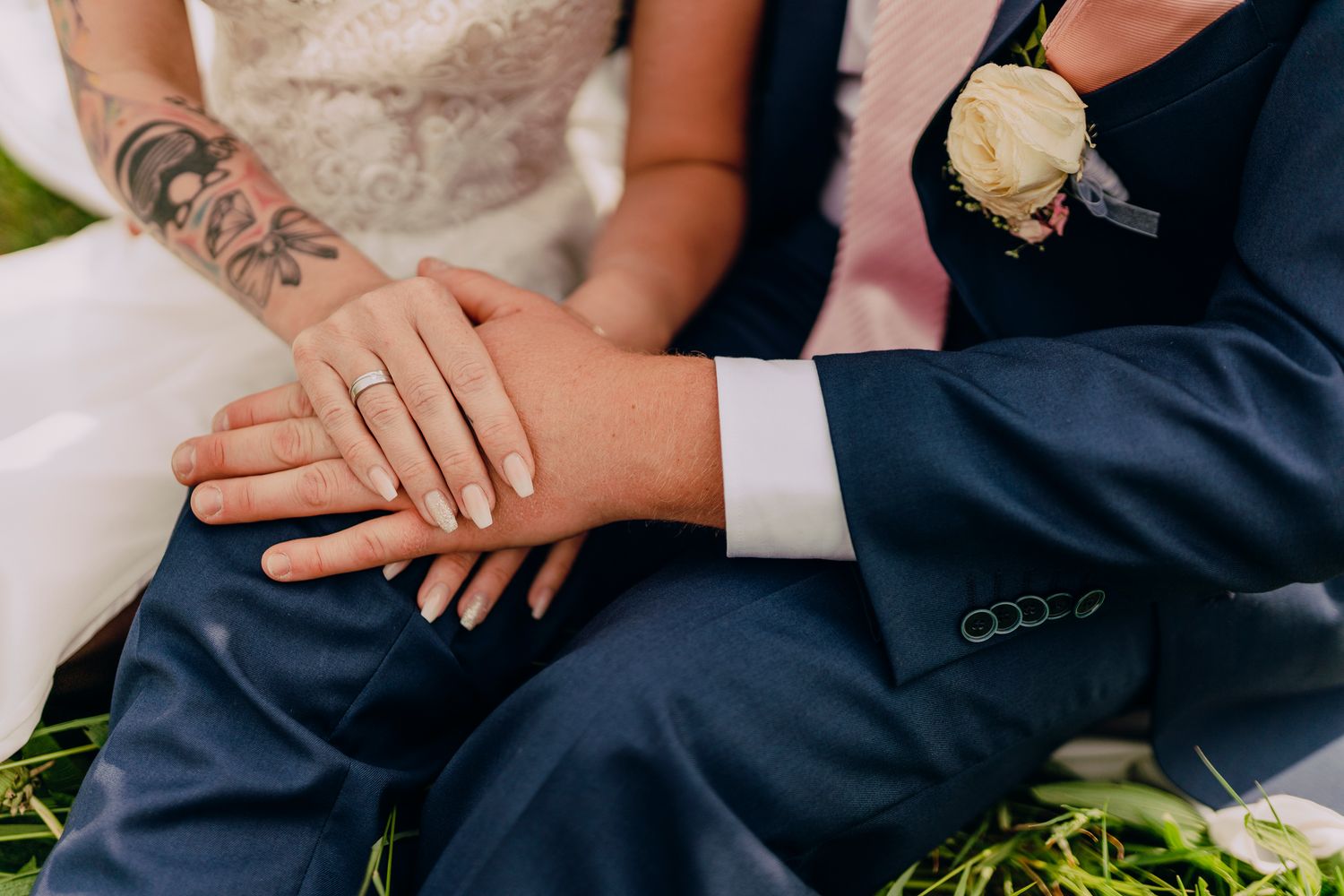 Close up of newlywed hands with wedding rings, white lace dress and navy suit, with floral boutonniere visible.