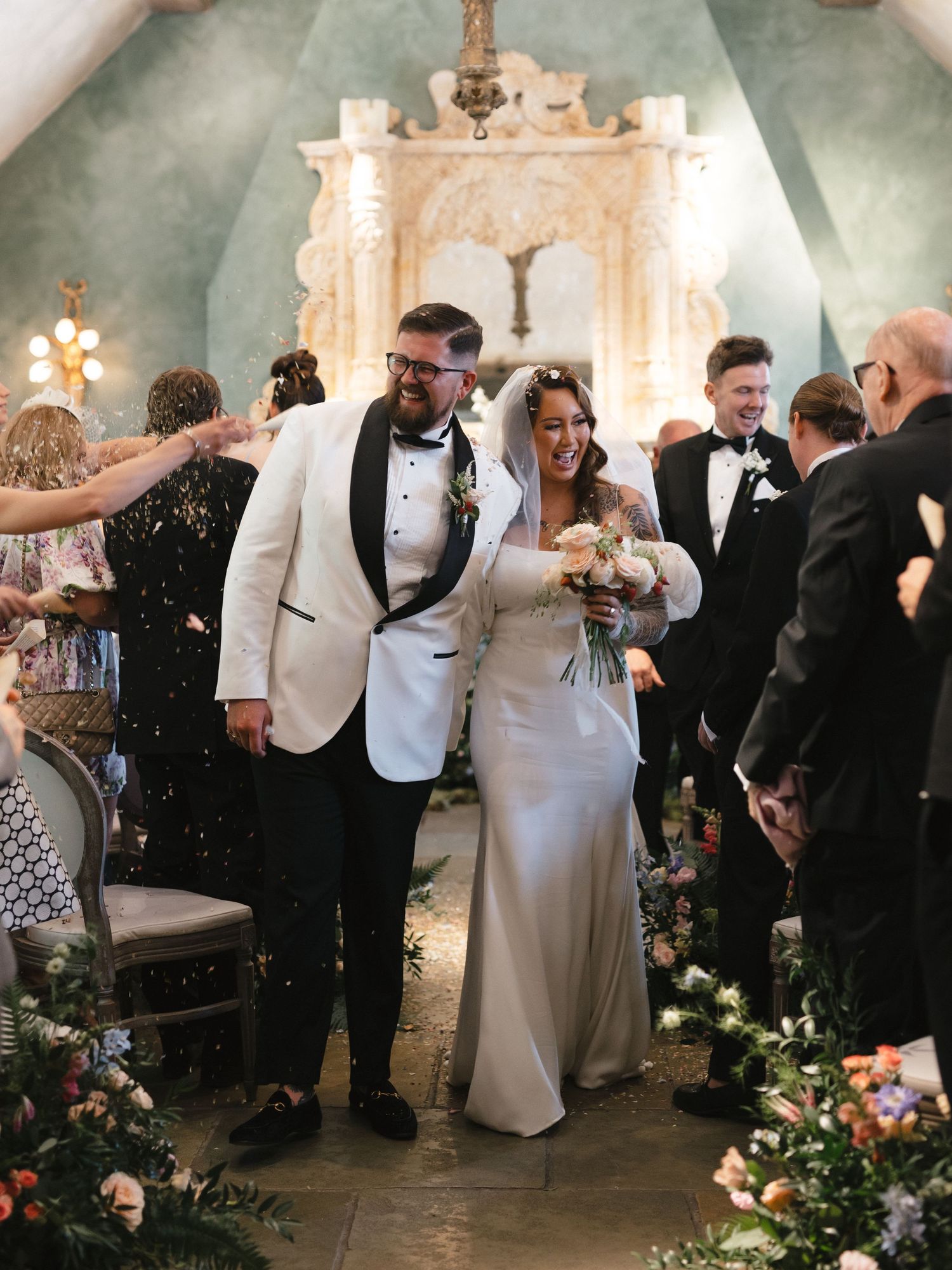 Newlyweds walk down aisle while guests throw confetti in ornate church venue with floral decorations.