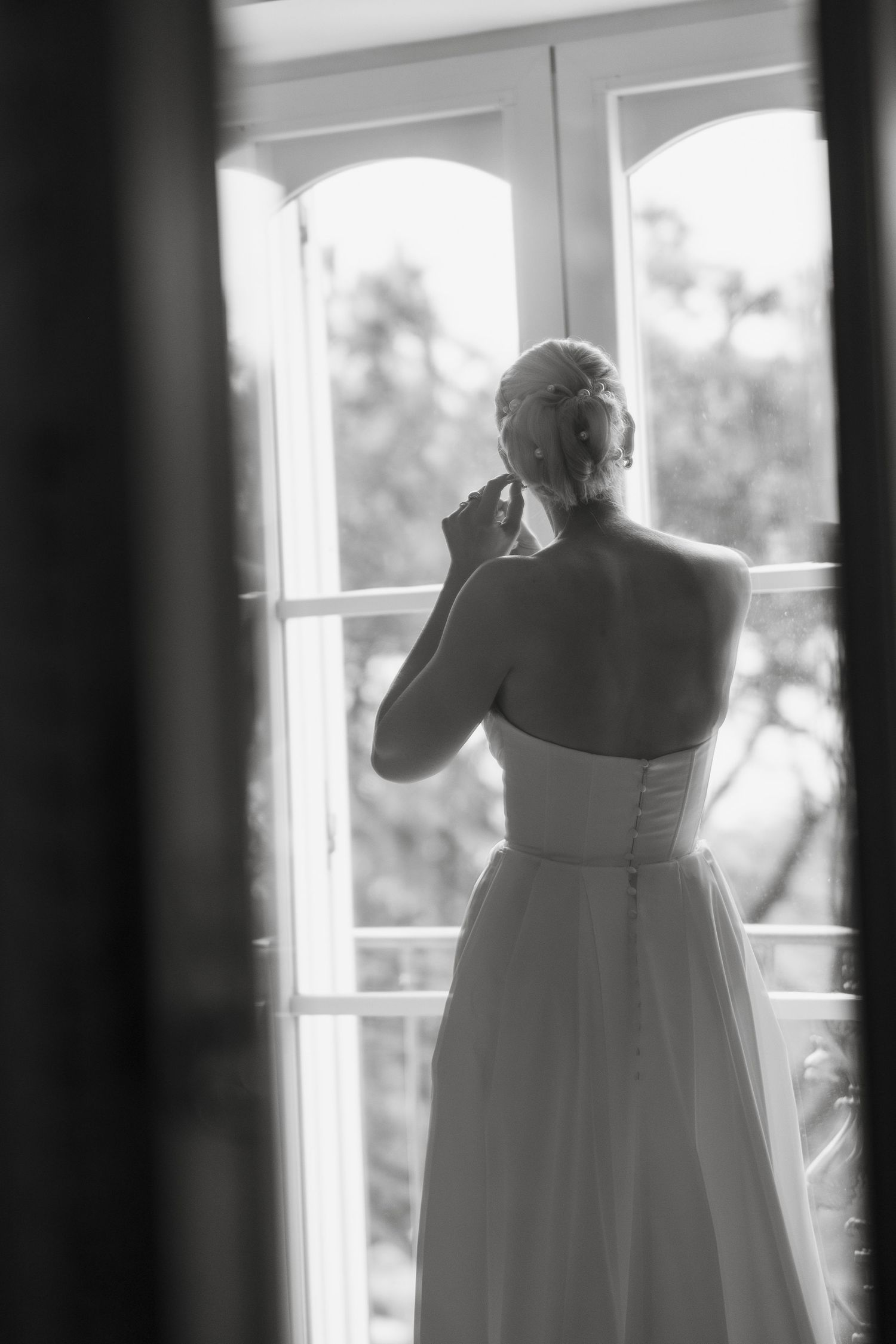 Black and white photo of someone in a wedding dress looking out a window in contemplative silhouette.