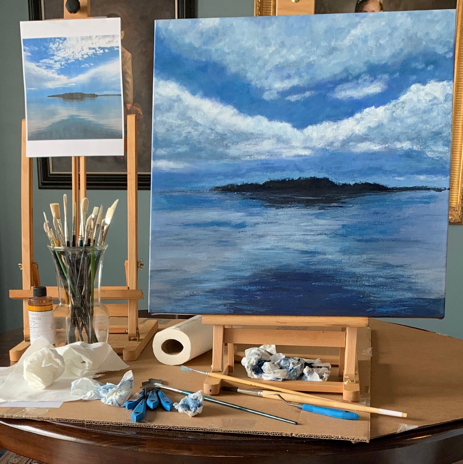 Coastal painting in progress on easel with art supplies, paints and brushes on a wooden table.