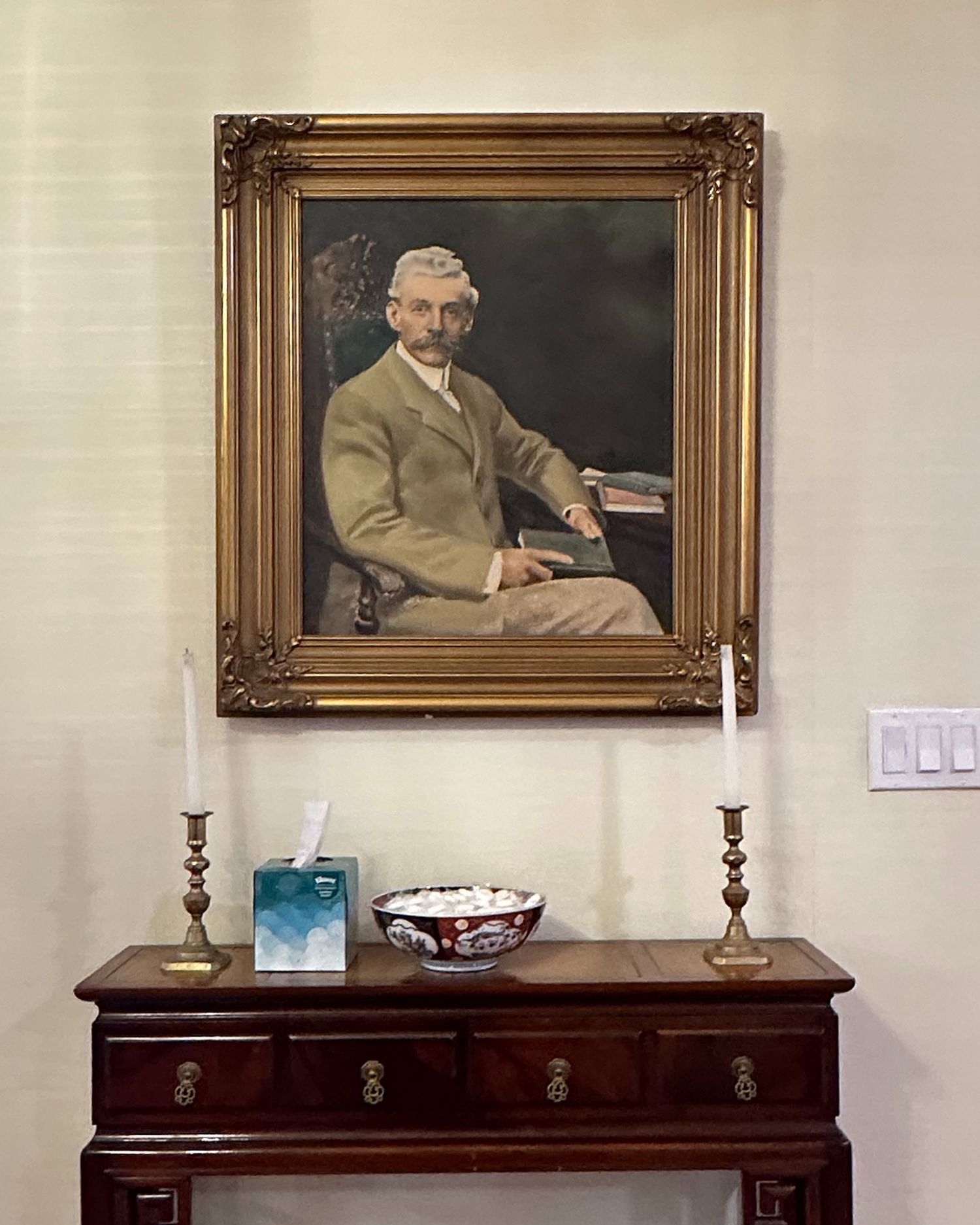 An ancestor's portrait in ornate gold frame hanging above antique wooden sideboard with candlesticks.