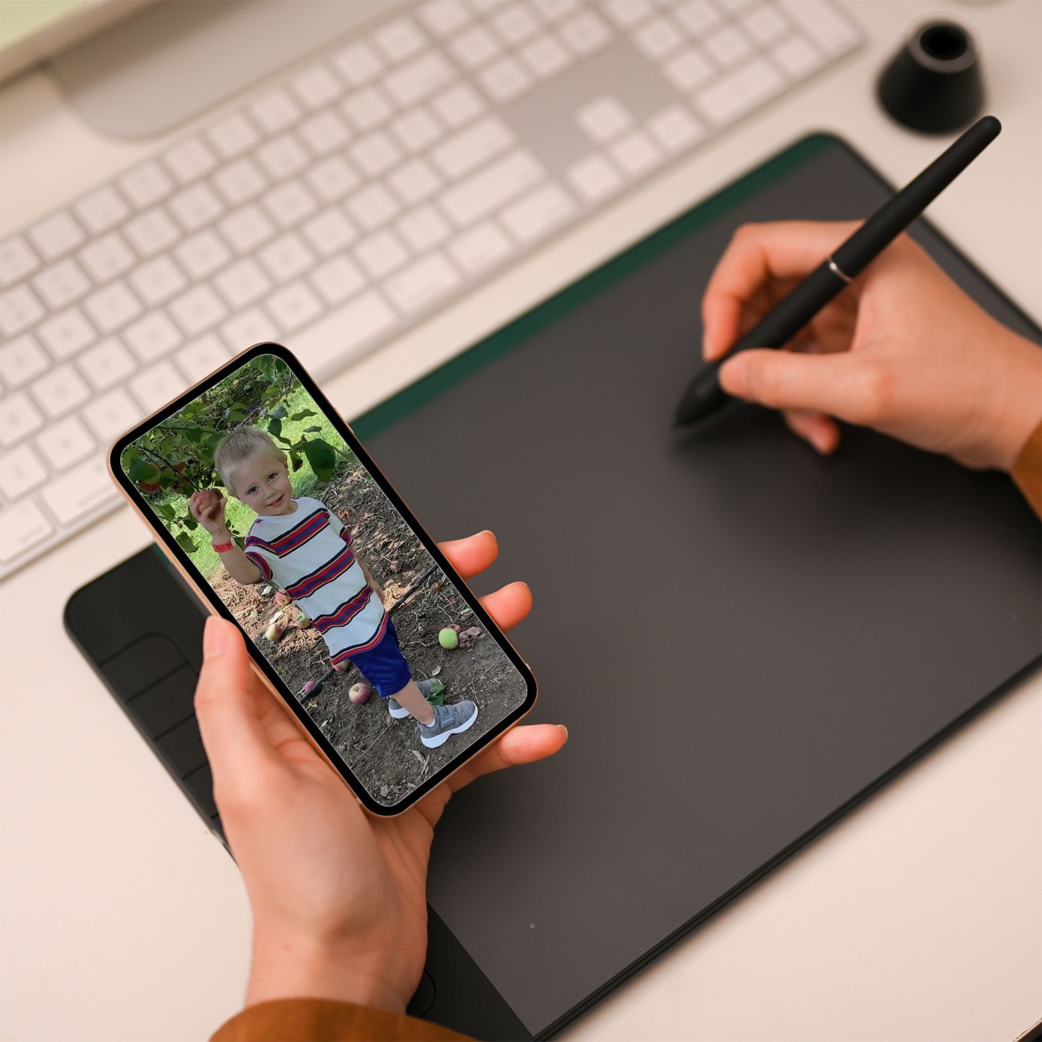 Artist working on a graphics tablet while holding a phone displaying an source photo of a boy.