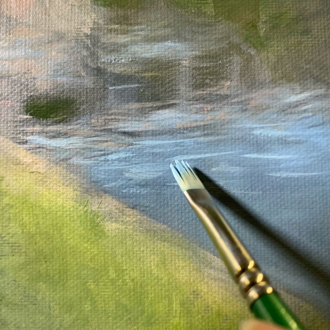 Close-up of an artist's paintbrush working on a blue and green landscape painting.
